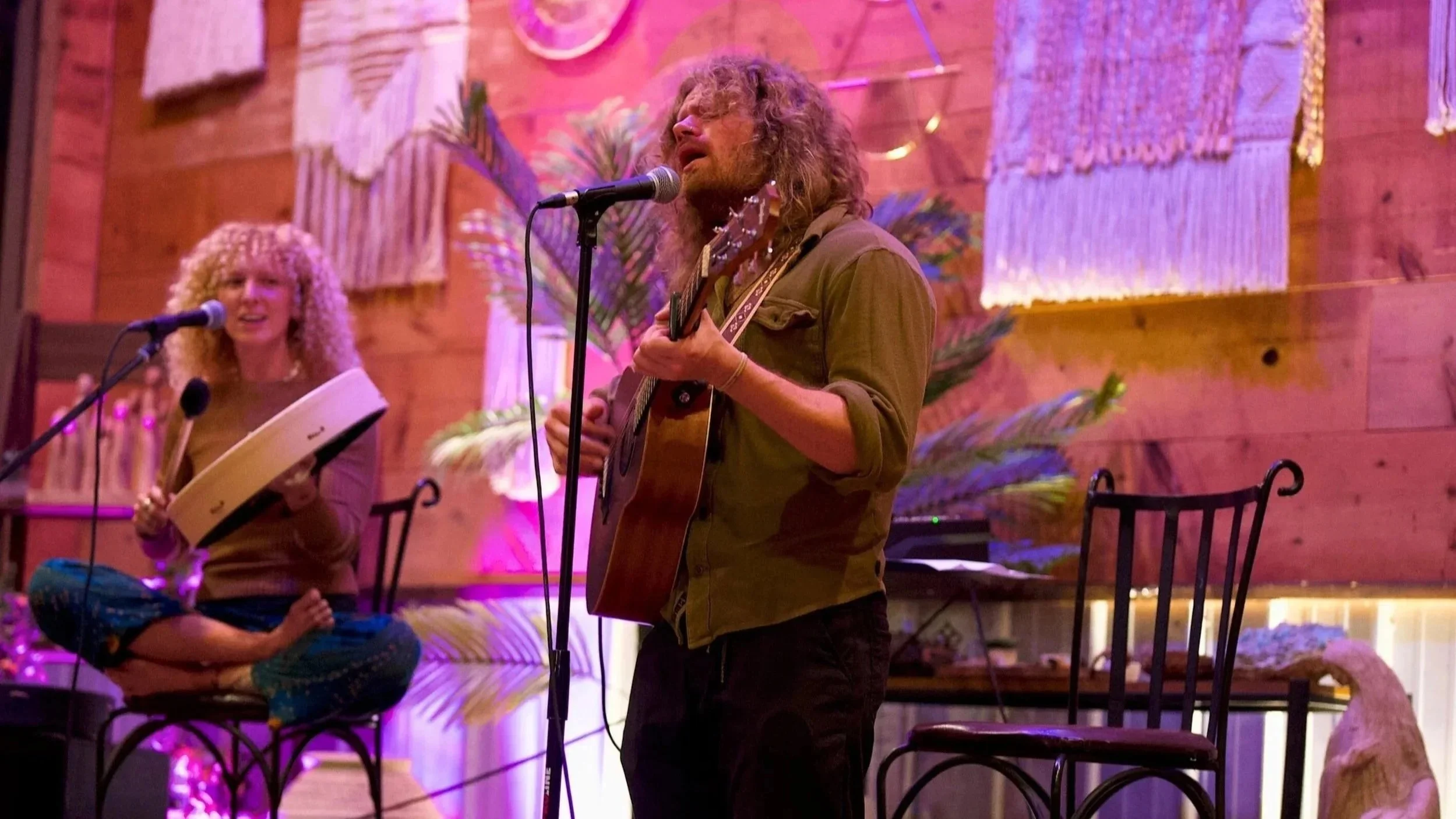 A man with long, curly hair and a beard playing an acoustic guitar and singing into a microphone. A woman with curly blonde hair playing a tambourine and singing into a microphone. They are in a decorated, cozy room with wooden walls, large plants, a