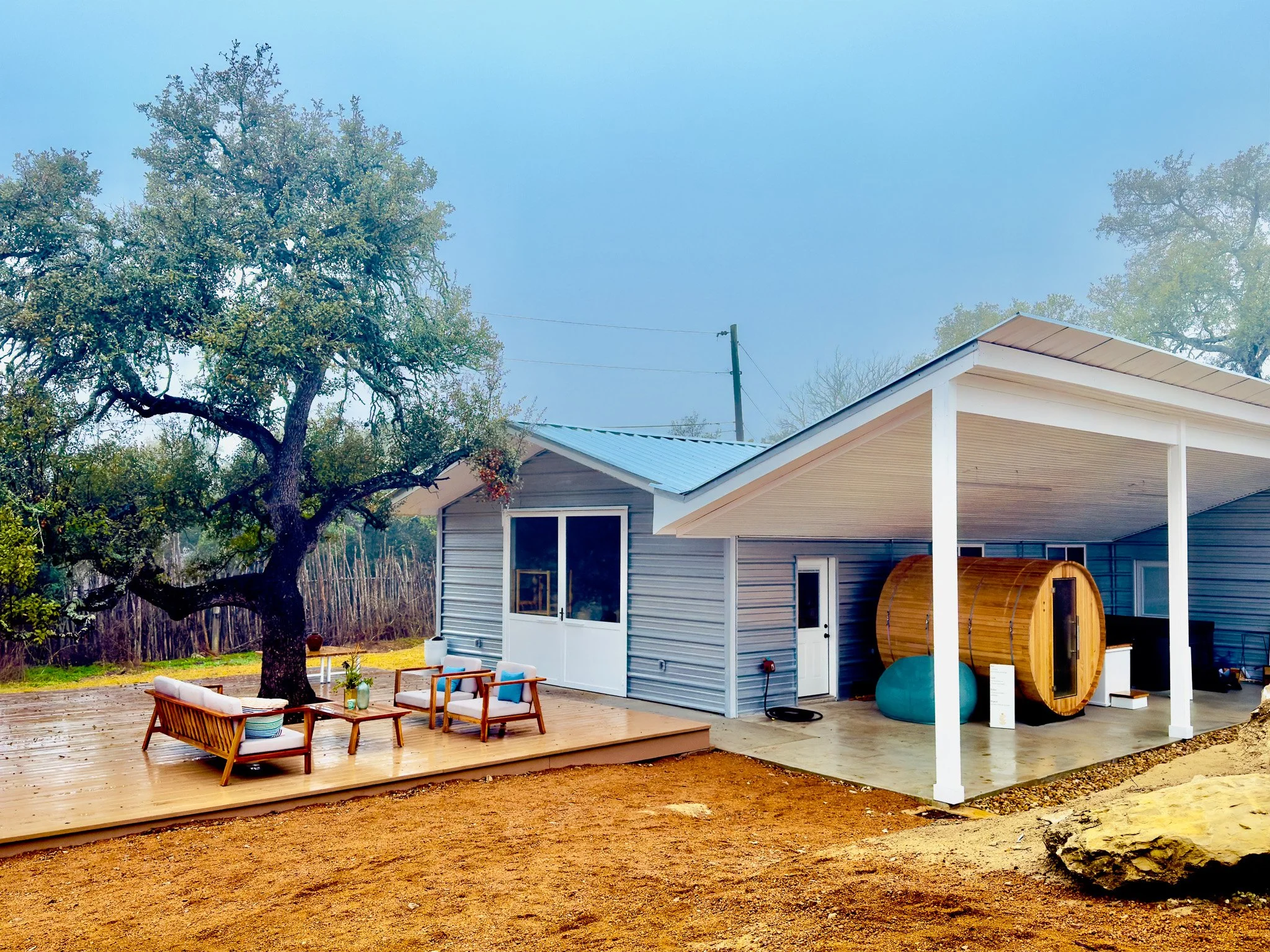 Backyard patio with outdoor seating under a large tree, adjacent to a house with a covered area featuring a wooden barrel sauna and outdoor furniture.