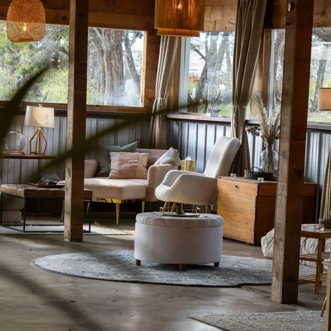 Cozy rustic living room with wood-paneled walls, large windows, beige sofa with pillows, white armchair on a round ottoman, side table with vase and dried flowers, and soft lighting.