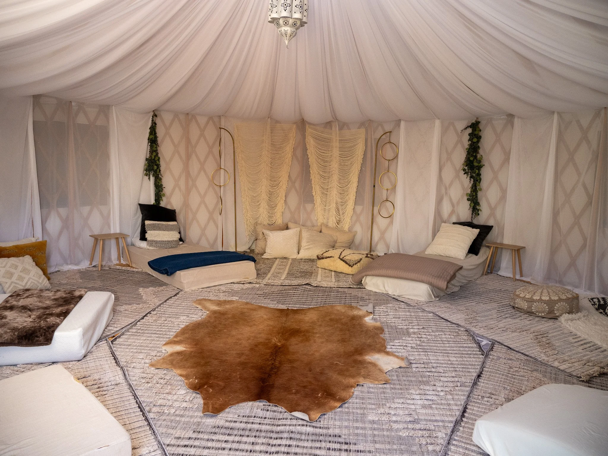 Interior of a bohemian-style tent with soft fabrics, cushions, and decorations on the walls, featuring a cowhide rug on a round area rug, surrounded by various cushions and seating options.