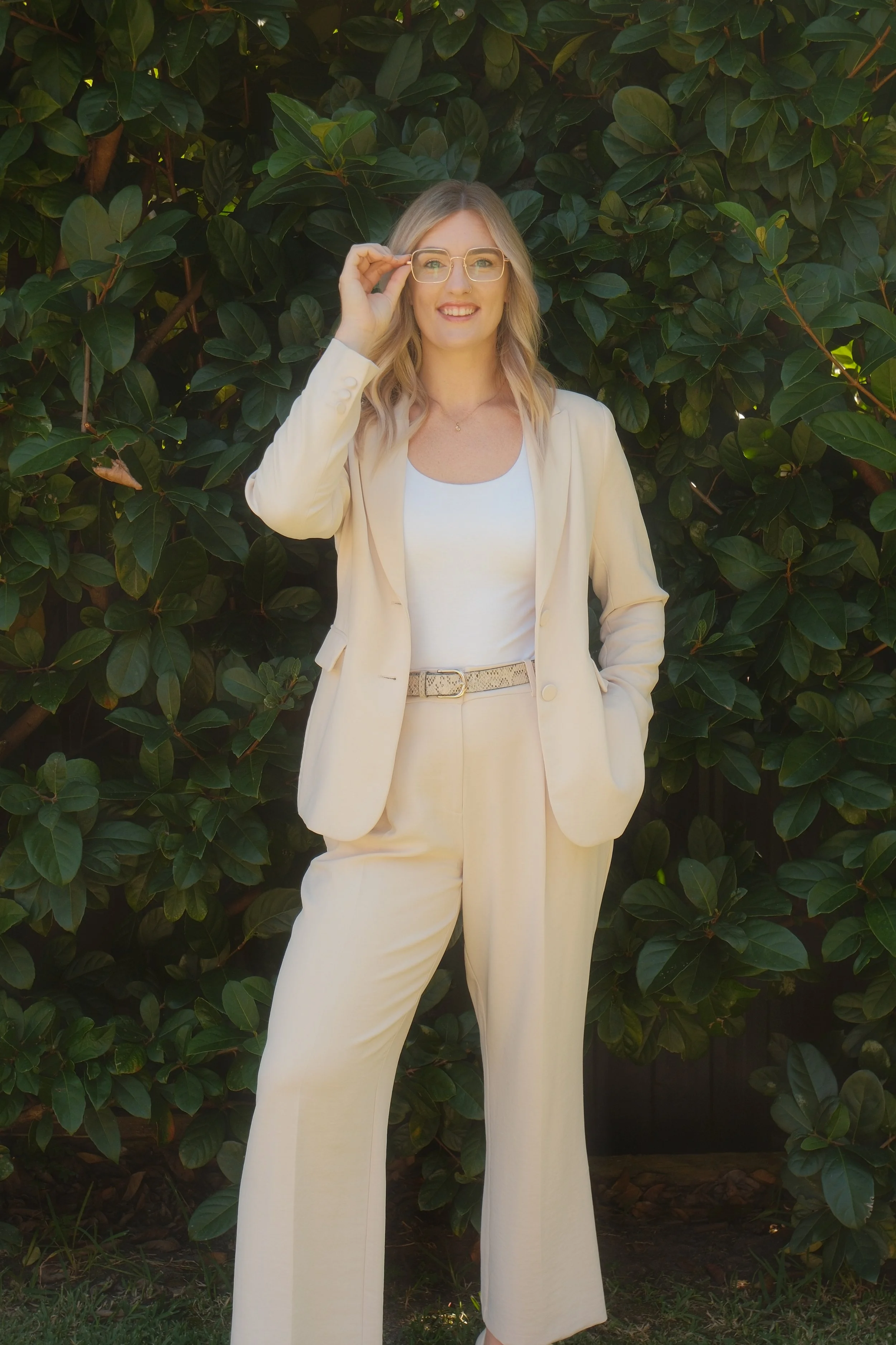 A woman wearing a cream-colored suit and glasses standing in front of leafy green bushes.
