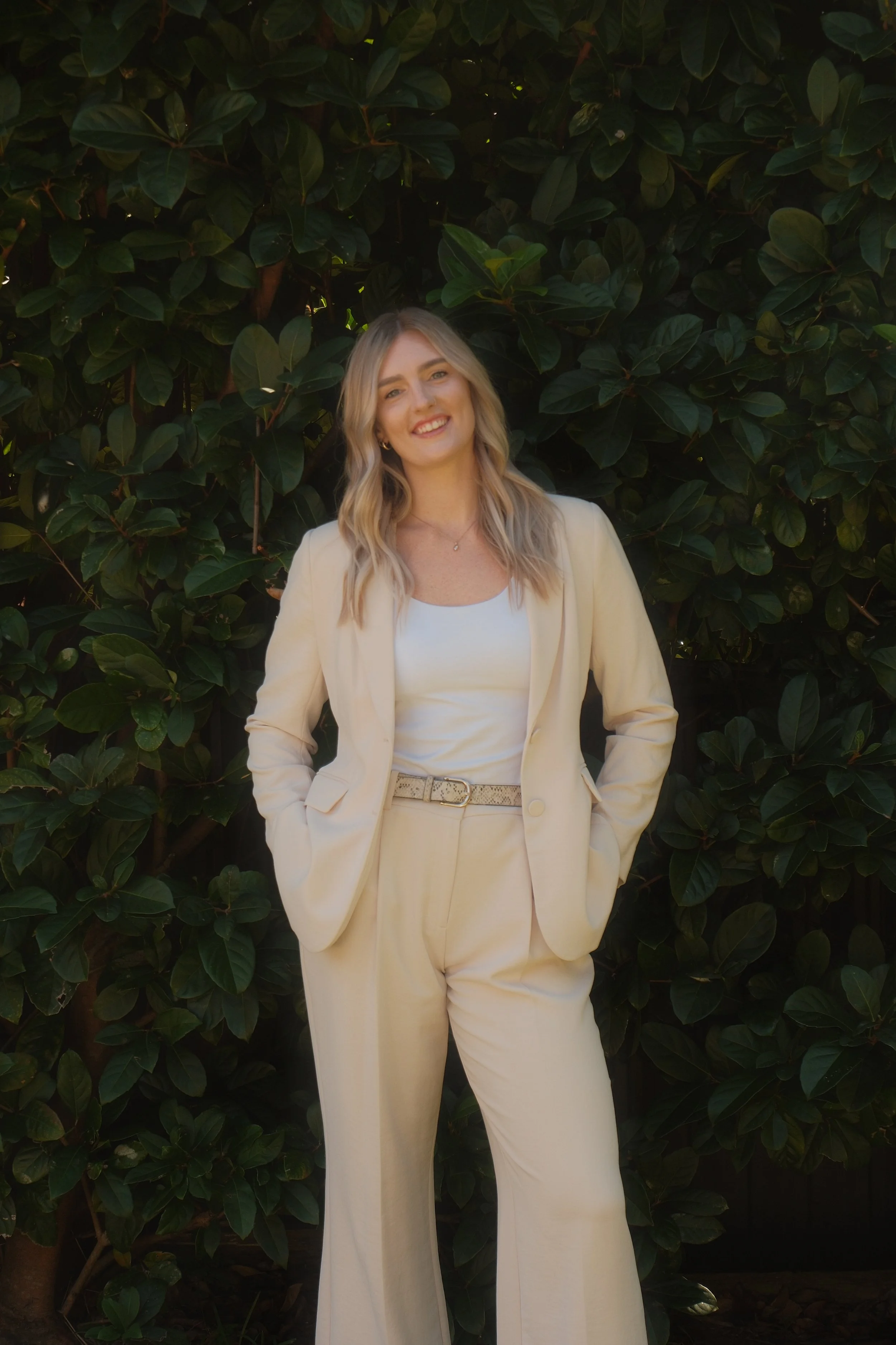 Young woman in a cream-colored suit standing outdoors in front of a leafy green background, smiling with hands in pockets.