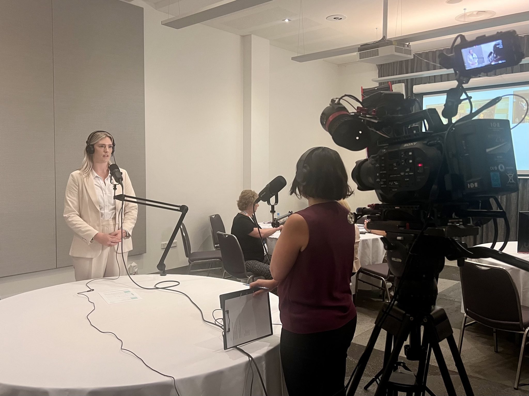 A woman in a white suit and headphones stands in front of a microphone, participating in an interview or podcast recording. A woman with a tank top and headphones operates a camera, recording the scene. Several chairs and a table with a laptop are in the background.