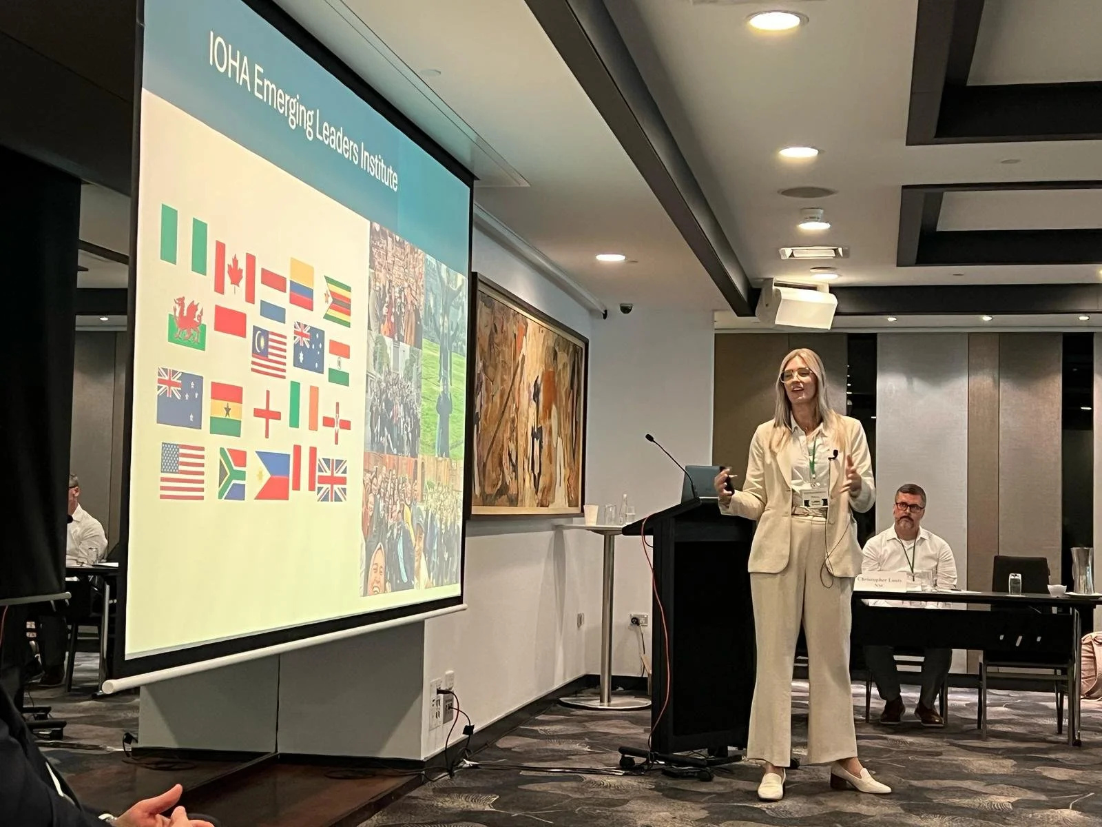 A woman in a cream-colored suit giving a presentation at a conference, standing next to a large screen displaying international flags and a crowd photo. Other attendees are seated at tables in the background.