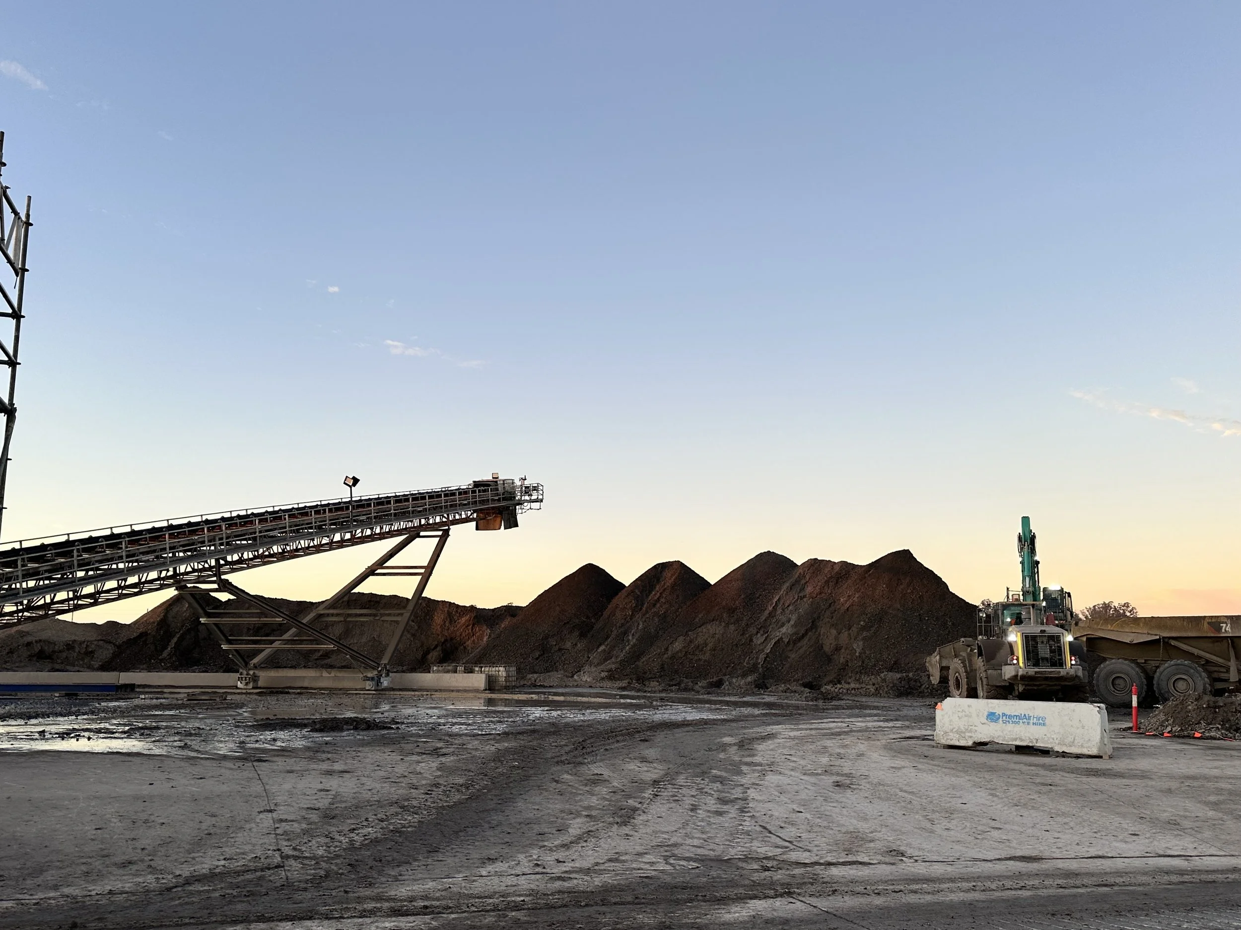 Construction site with a large conveyor system and piles of dirt or gravel, a dump truck, and construction barriers at sunset.