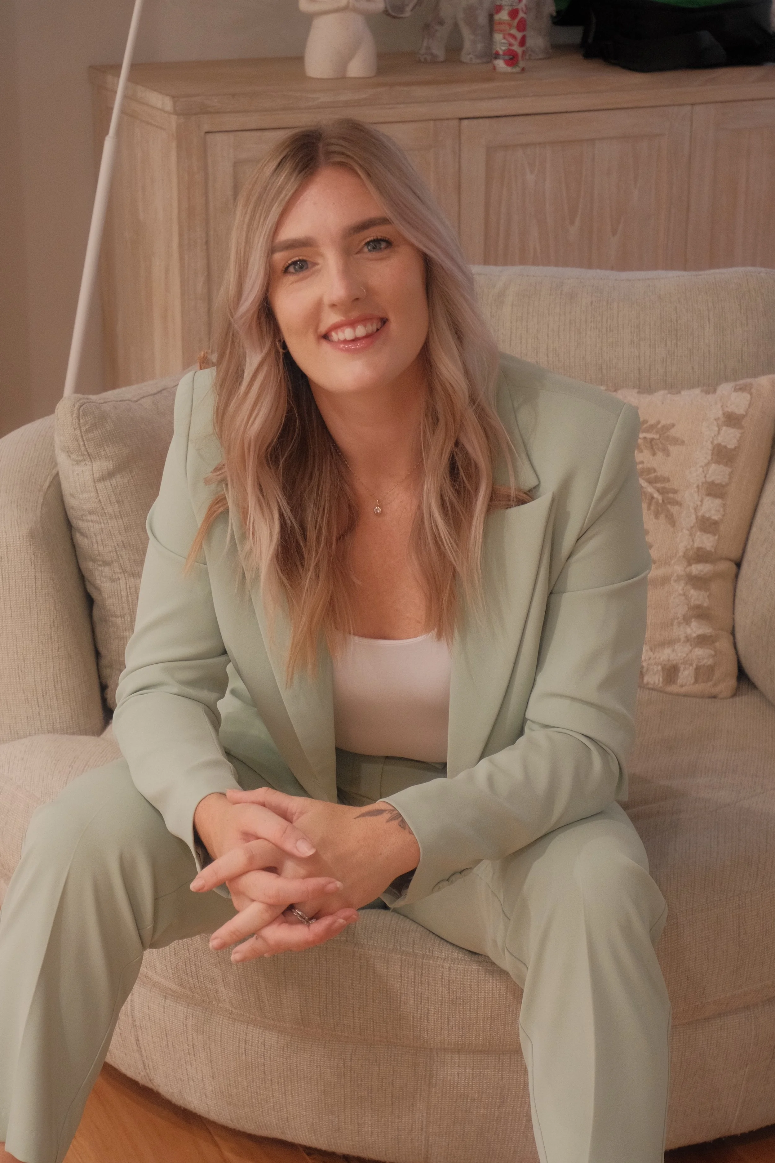 A smiling young woman with long, wavy blonde hair wearing a light green blazer and matching pants, sitting on a beige sofa with a beige cushion, in a cozy living room.