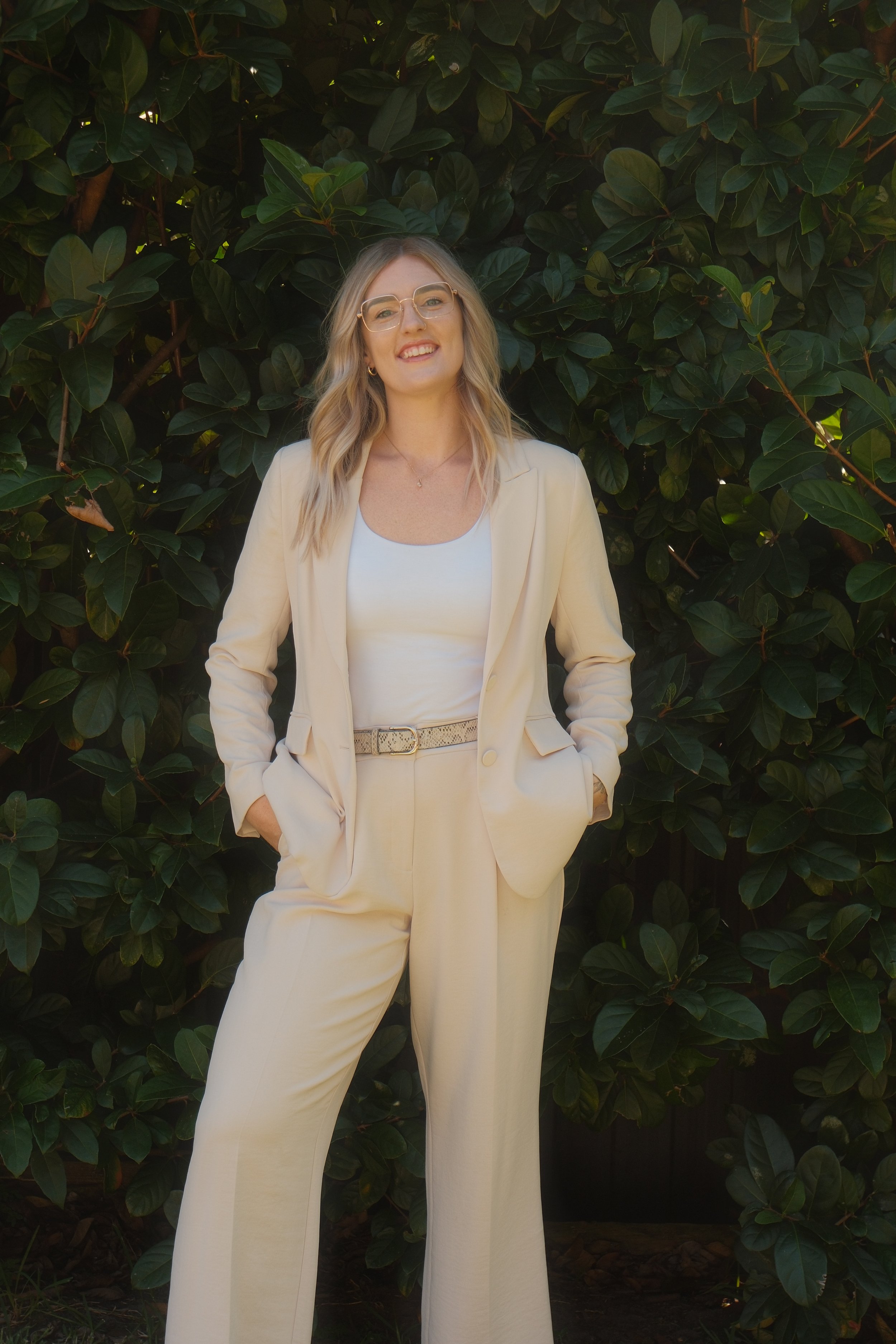 A woman with blonde hair and glasses wearing a cream-colored suit and white top, standing outdoors with greenery in the background.