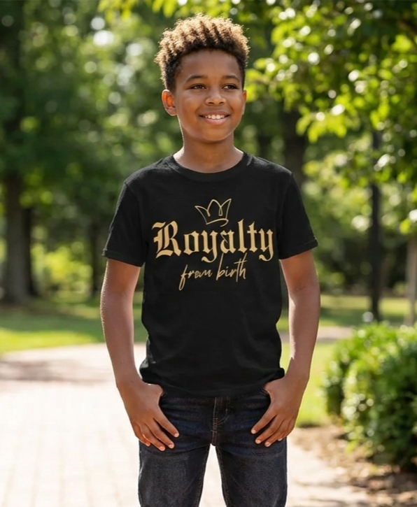 Smiling boy standing outside in a park with trees and a pathway in the background, wearing a black t-shirt that says "Royalty from birth" with a crown graphic.