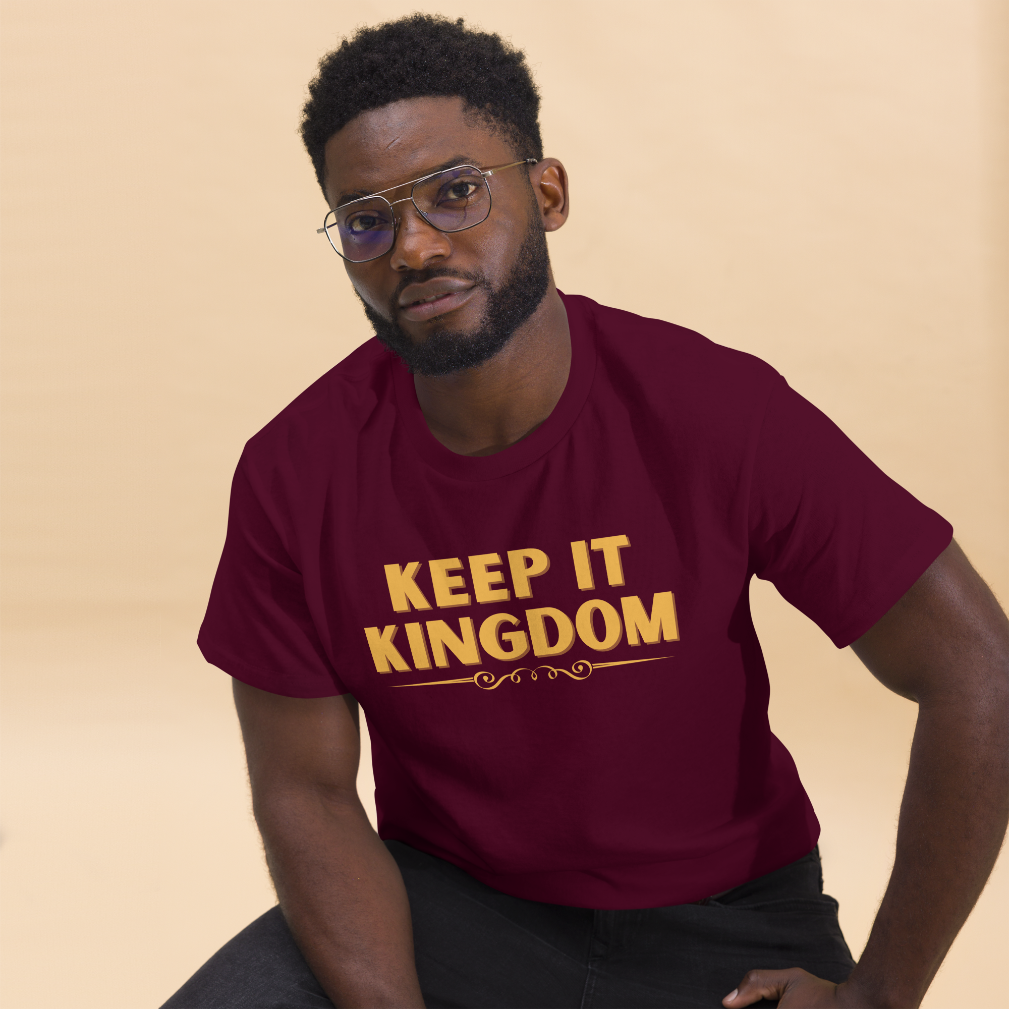 A man with glasses and a beard wearing a maroon T-shirt that says 'KEEP IT KINGDOM' in gold letters, sitting against a beige background.