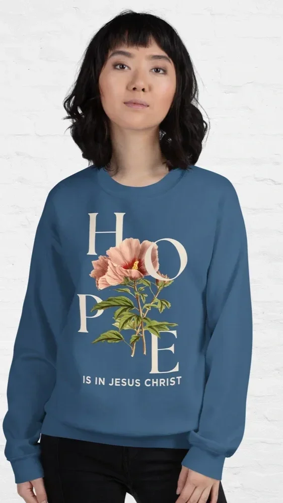 Woman with shoulder-length black hair wearing a blue sweatshirt with the word 'HOPE' and a pink flower graphic, standing against a white brick background.