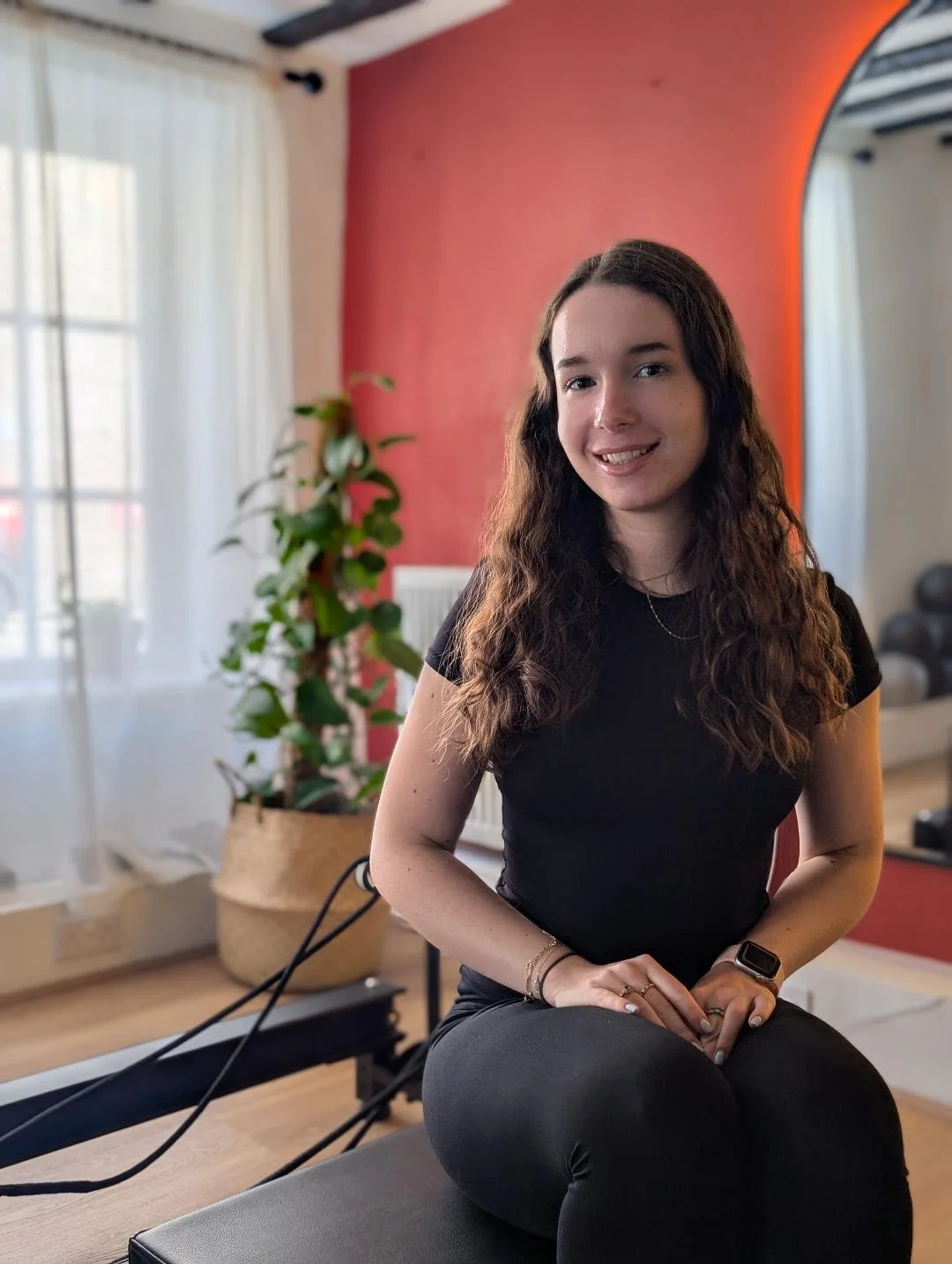 A young woman with long wavy brown hair, wearing a black t-shirt and black pants, sitting on a gym bench in a fitness room, smiling at the camera. In the background, there is a red wall, a potted plant, and gym equipment.