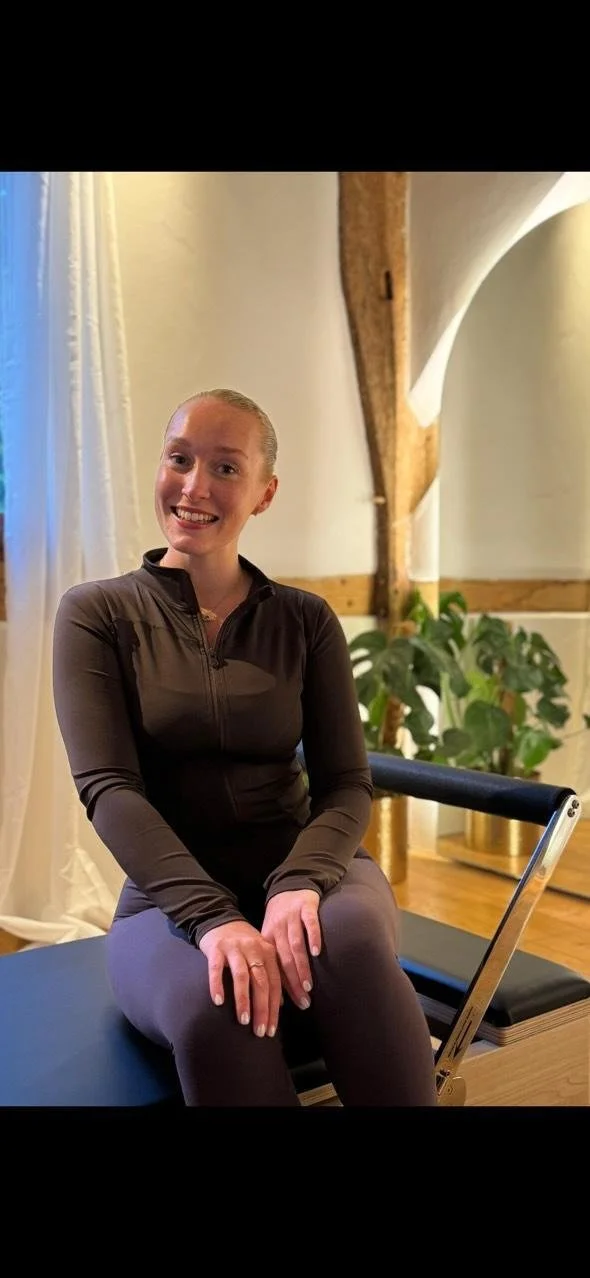 A woman sitting on a Pilates reformer machine in a room with wooden beams and green plants, smiling at the camera.