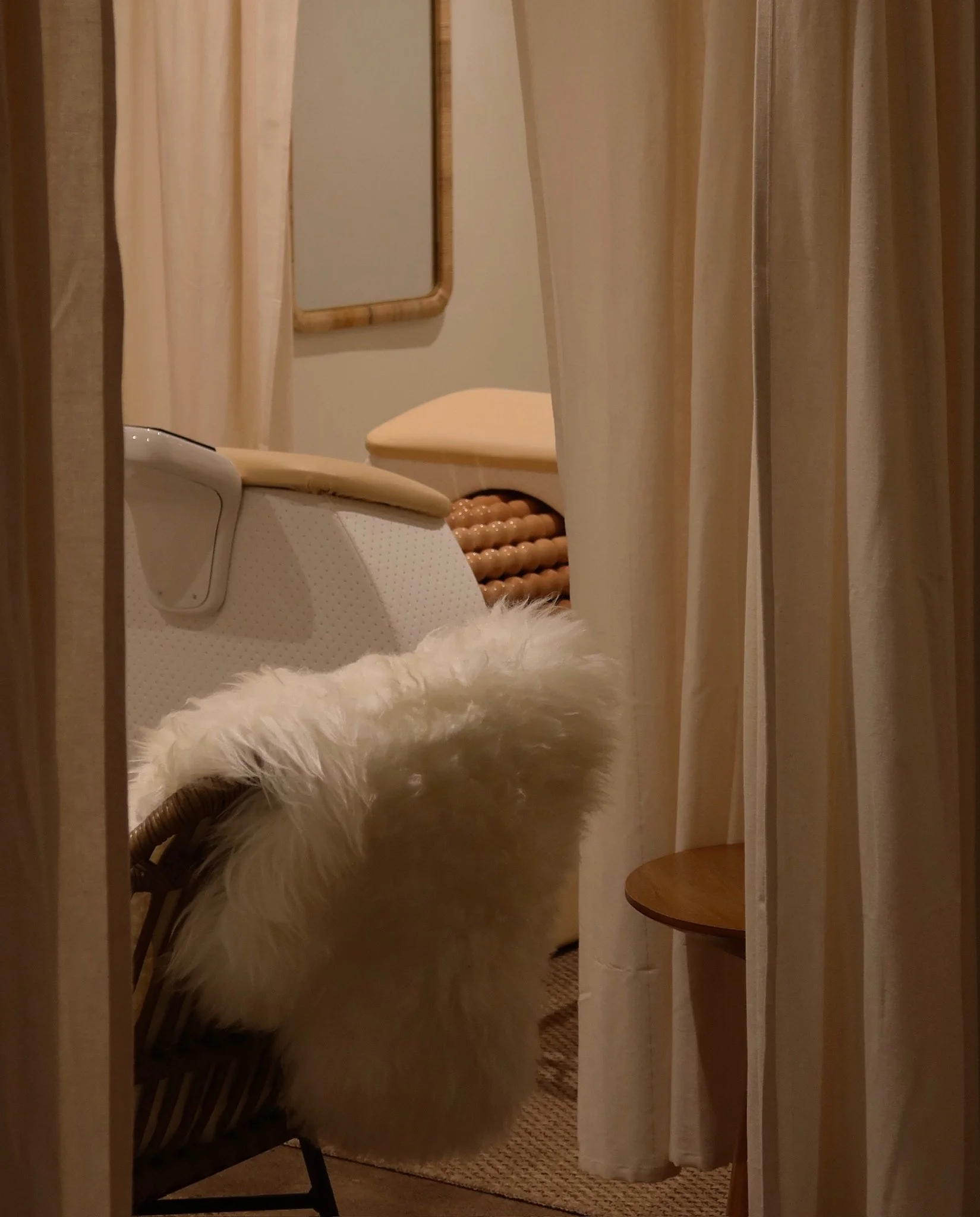 View through a curtain into a room with a cream-colored massage or treatment table, a fluffy white chair, a mirror, and a wooden stool.