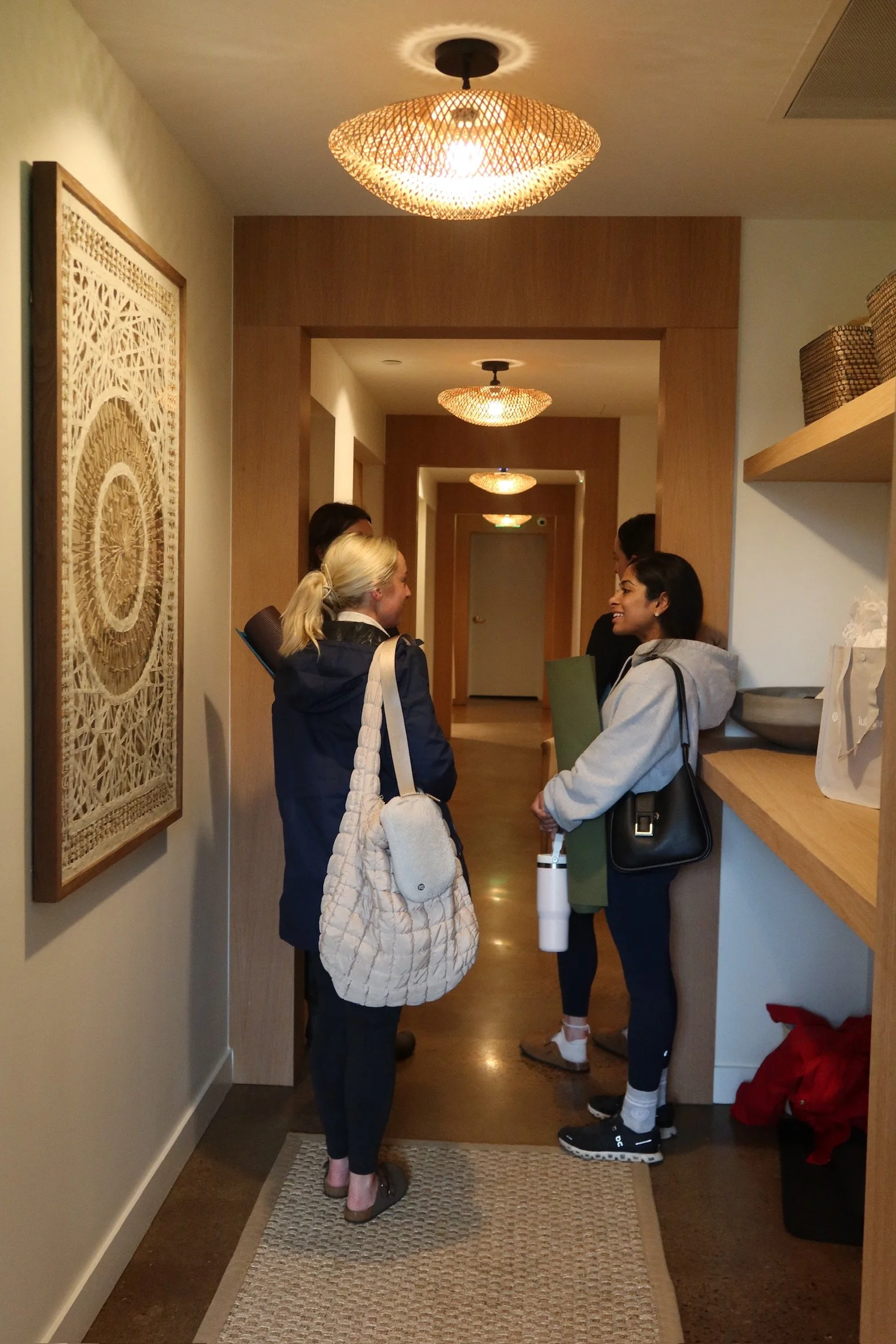 Group of five women talking in a hallway of a modern building, with warm lighting, wooden accents, and decorative wall art.