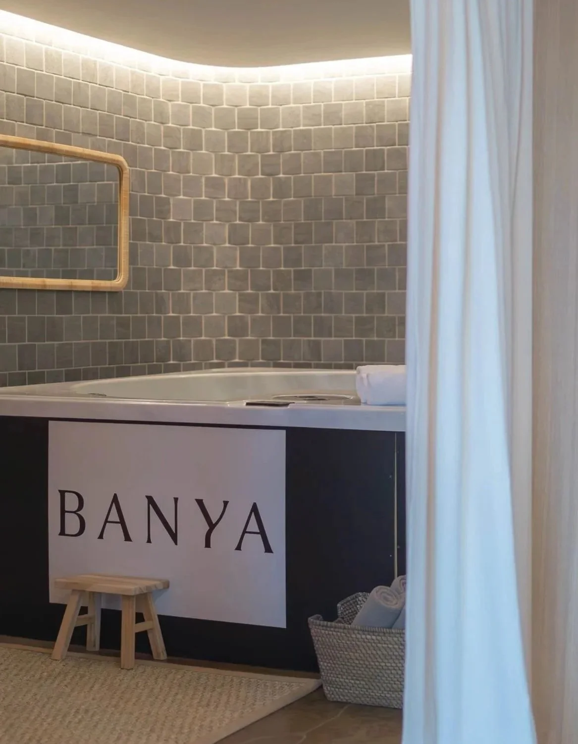 A bathroom with a jacuzzi tub, a mirror, and a sign that says 'BANYA' on a black panel, with a curtain on the right side.