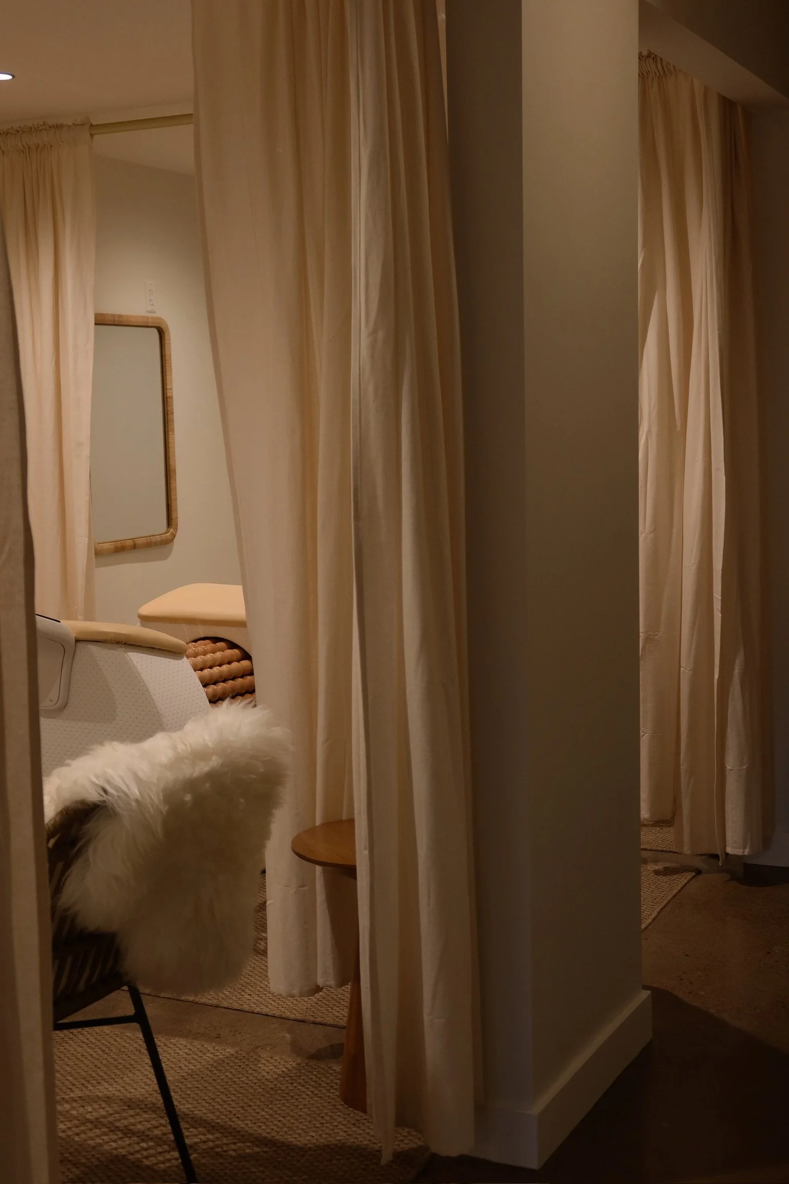 A cozy room with cream-colored curtains, a sheepskin-covered chair, a small wooden side table, and a mirror on the wall.