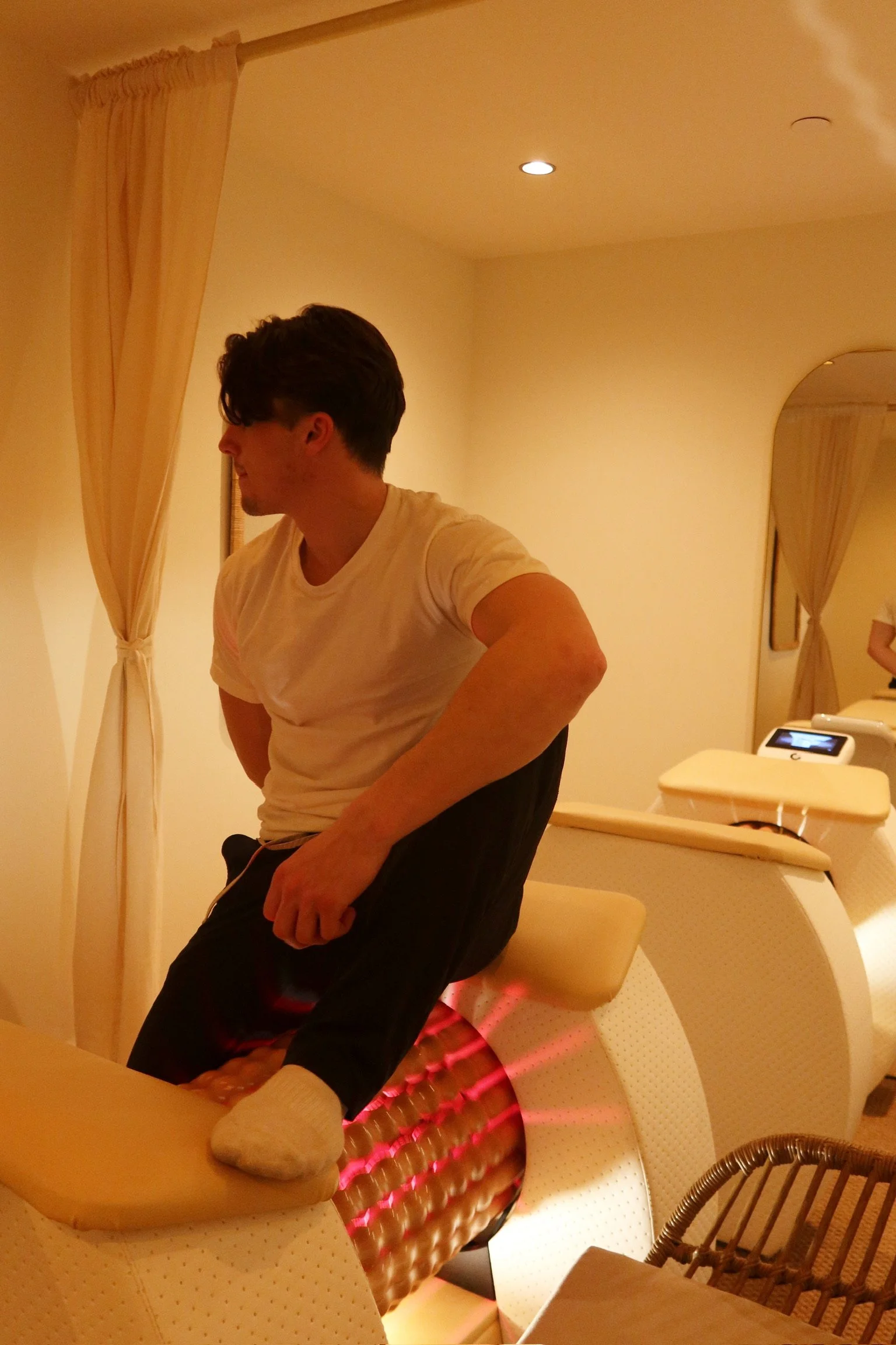 A man in a white shirt sitting on a massage chair, in a room with cream-colored walls and curtains, and a mirror reflecting part of the room and another person.