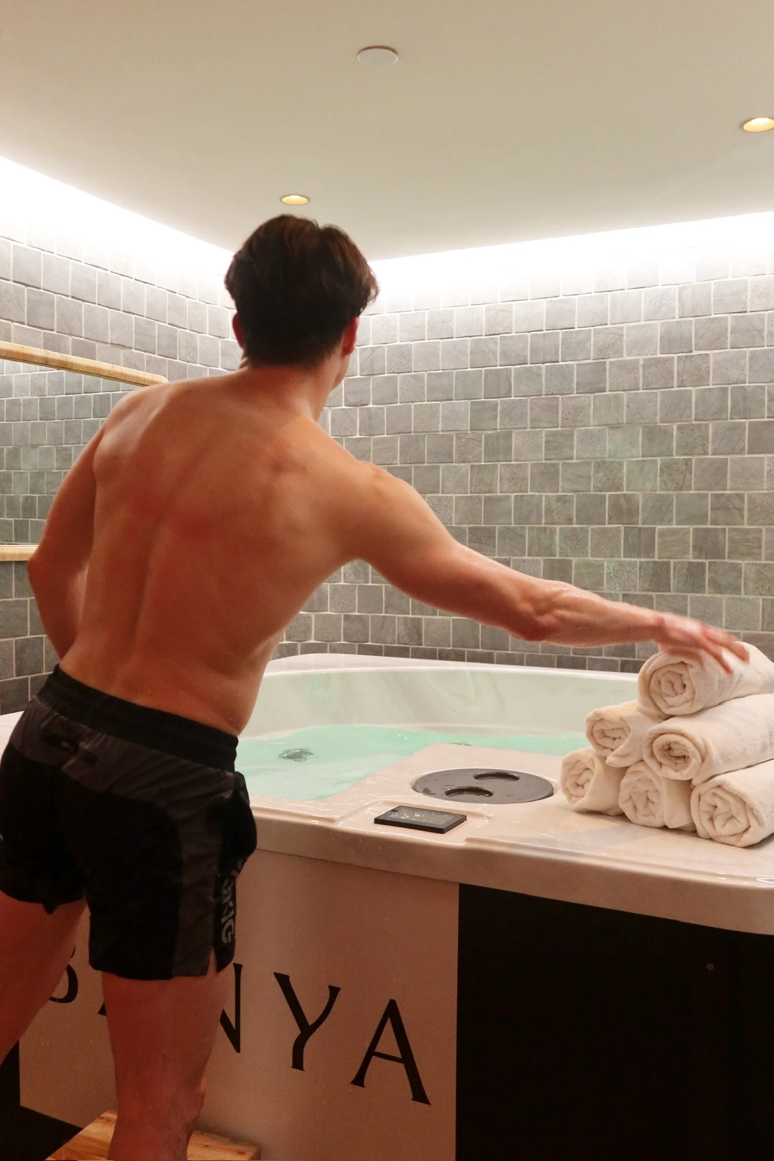 A man in black shorts is reaching to grab a rolled towel in a spa or wellness center with gray brick walls and a hot tub.