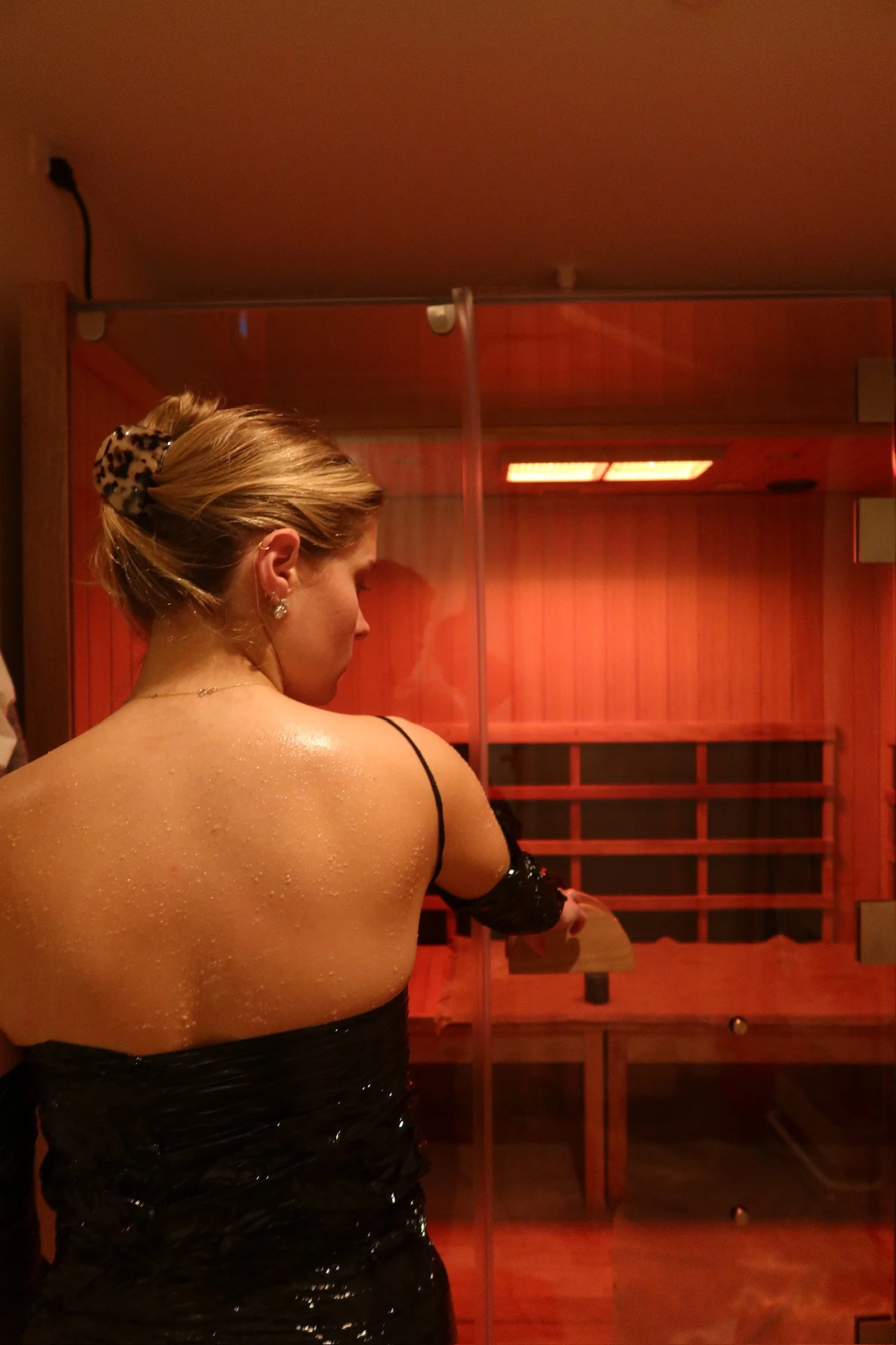 A woman in a black dress with wet skin standing in front of a sauna. She has blonde hair tied back with a leopard print scrunchie and wears earrings.