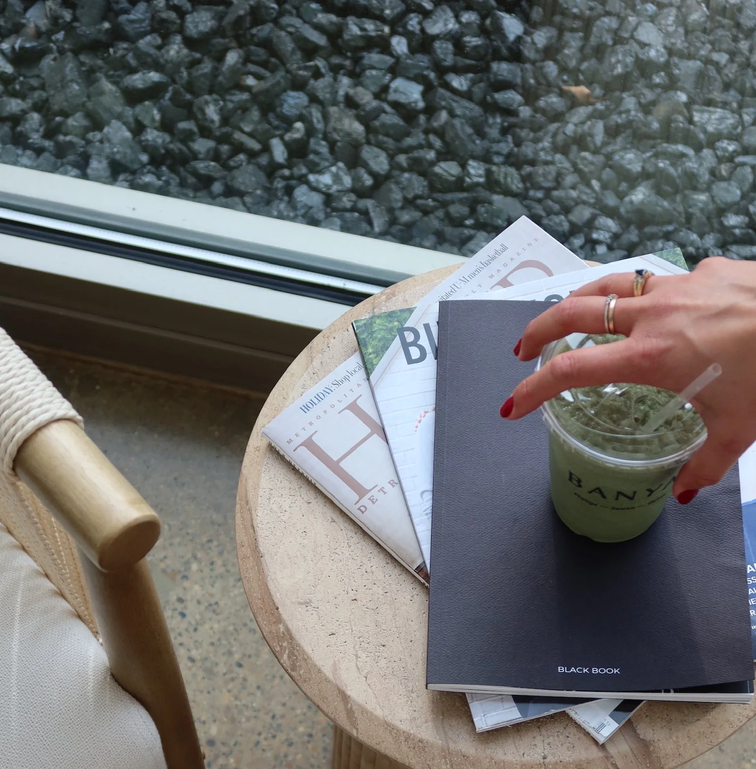 A small round table with magazines, a black notebook, and a hand holding a plastic cup with a green iced drink, located next to a window with rocks outside.