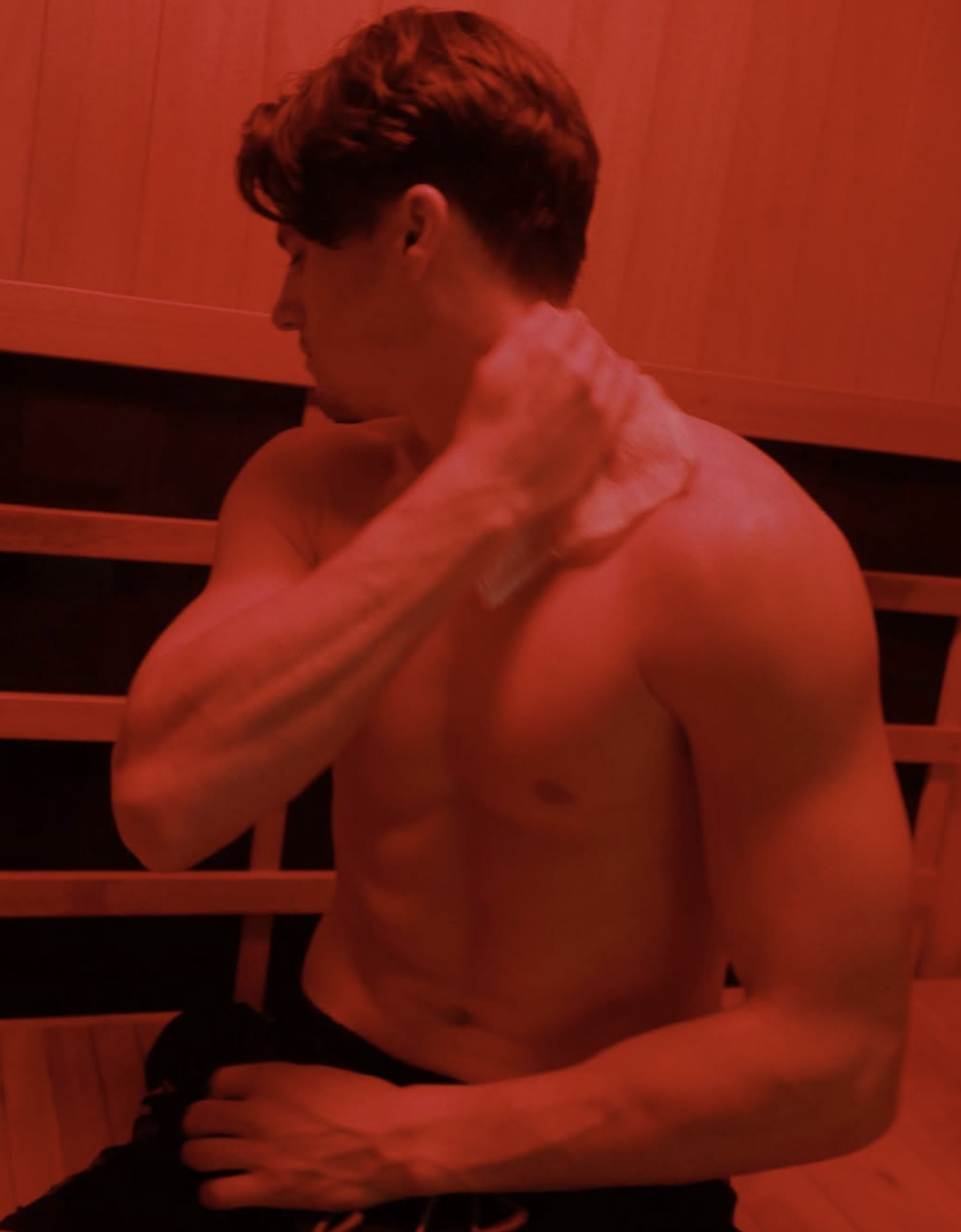 A shirtless young man with brown hair is sitting in a dimly lit room with warm lighting, touching his neck with his right hand and looking downward.