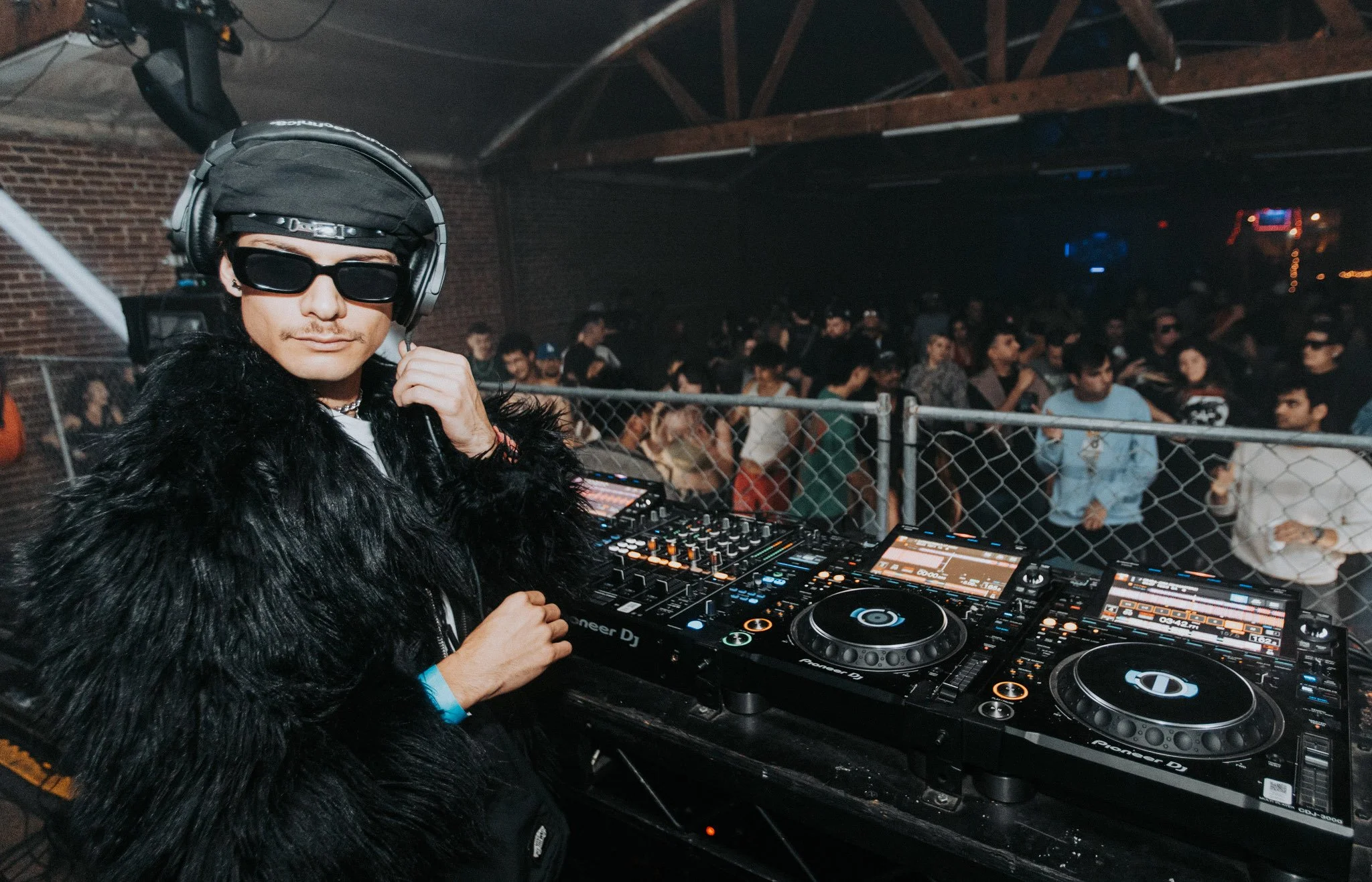 DJ wearing sunglasses, headphones, and a black faux fur jacket, standing behind a DJ booth in a nightclub with a crowd of people dancing in the background.