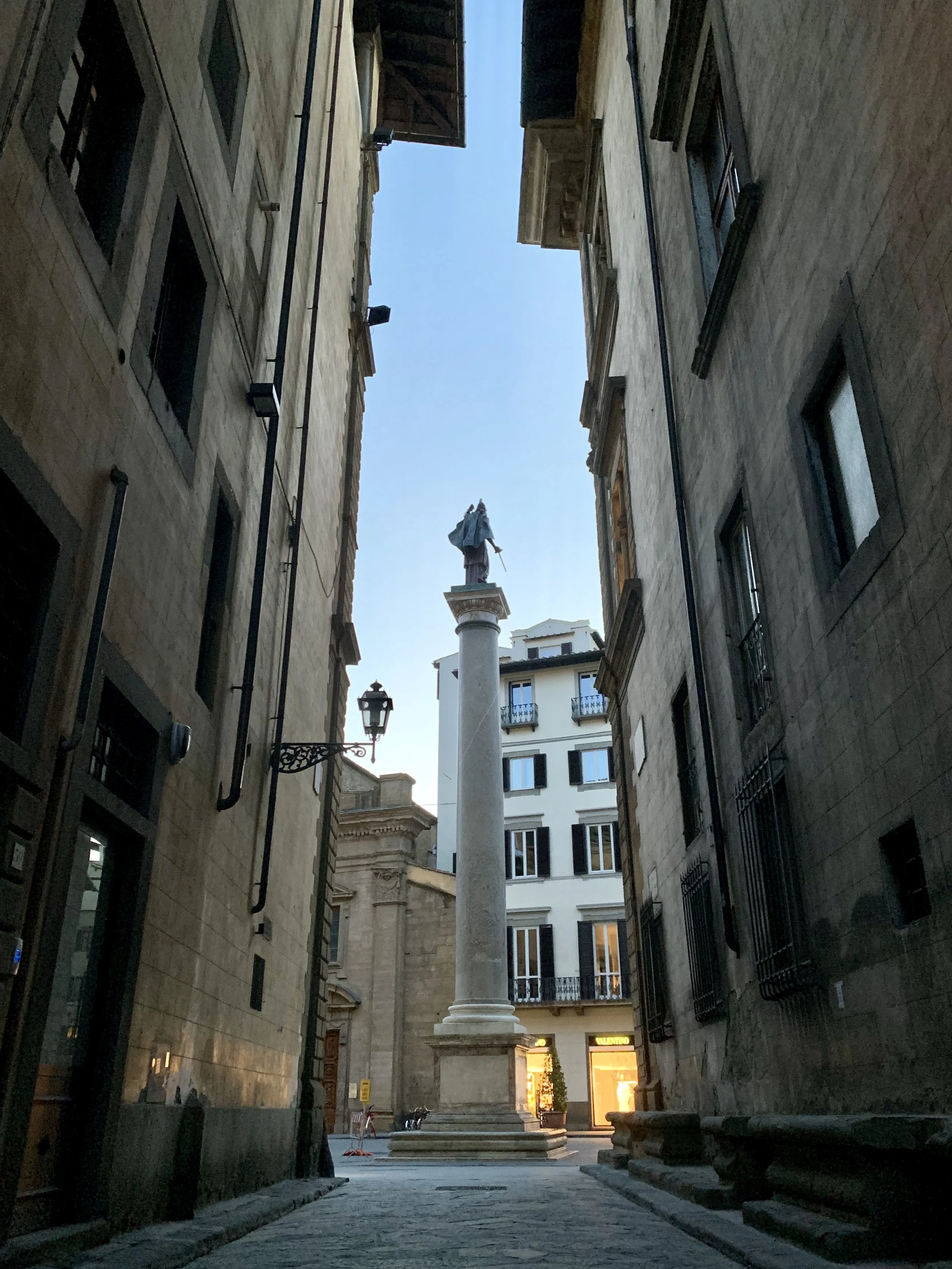 Veduta di una stretta via in centro città con un monumento con statua e colonne al centro, tra edifici storici, con il cielo visibile sopra.