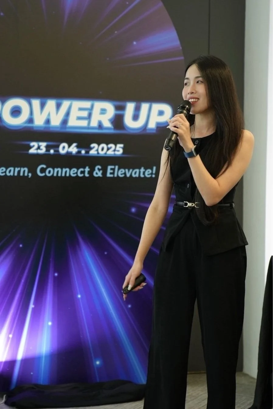 A woman in black outfit speaking into a microphone in front of a digital presentation screen with the words 'Power Up' and the date 23.04.2025, at a conference or seminar.