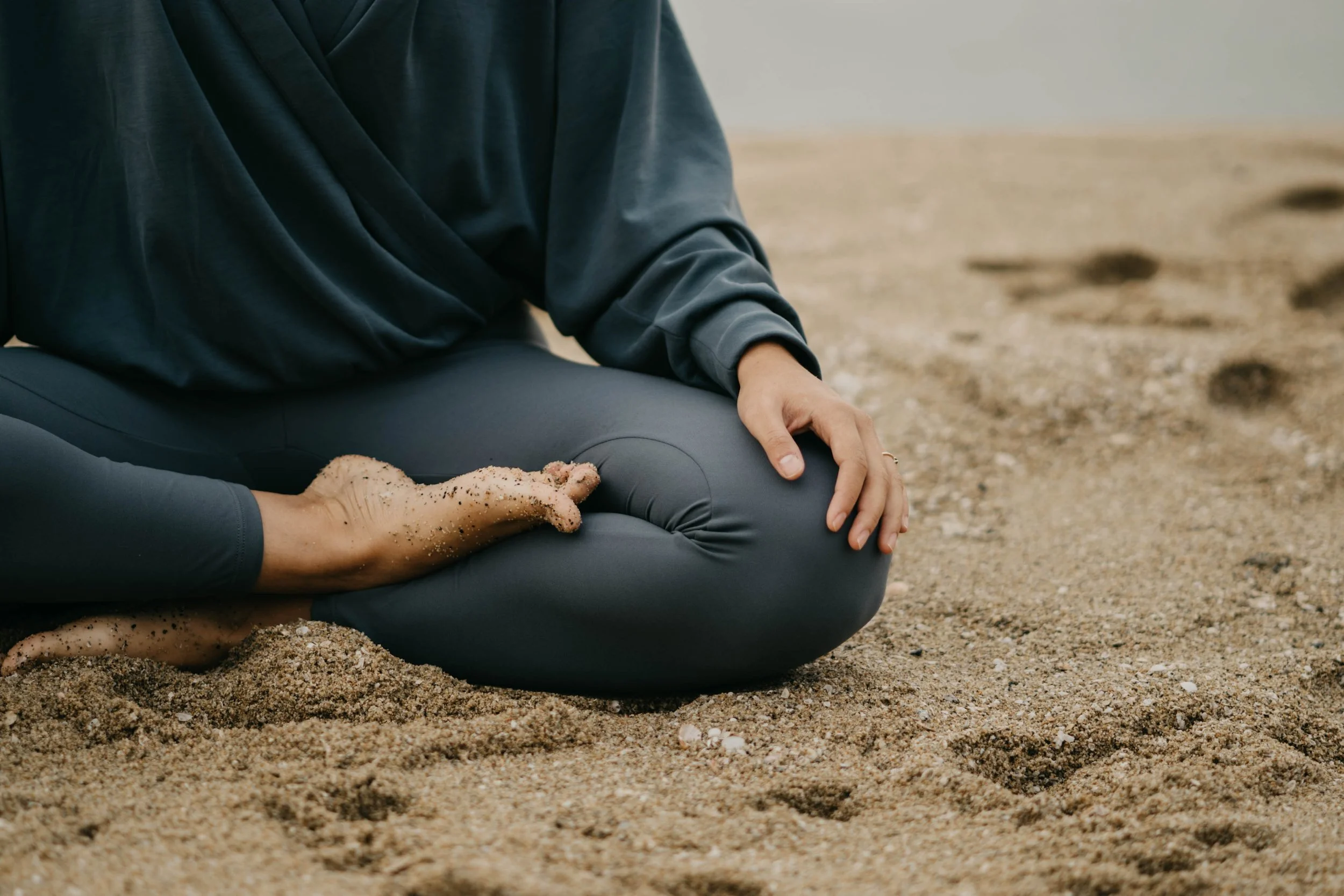 Persona en posición de meditación en la arena, usando ropa deportiva gris oscuro, con arena en las manos.