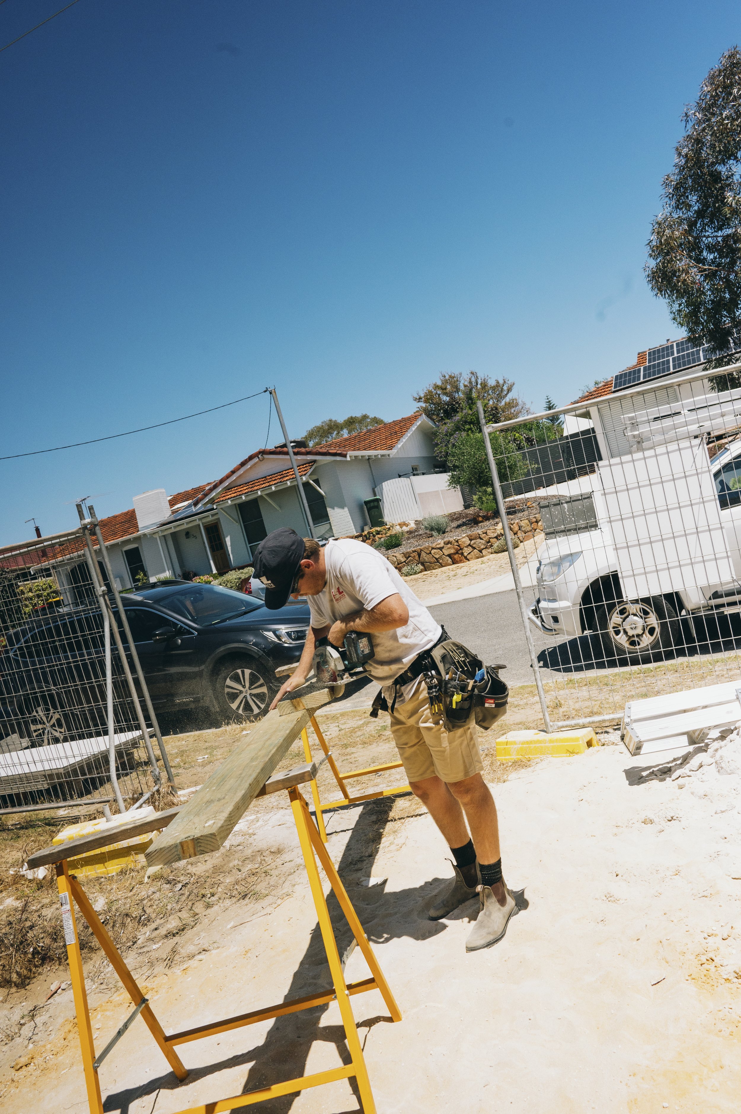 A construction worker using a power saw to cut a wooden board on a sawhorse at a construction site, with houses and vehicles in the background.
