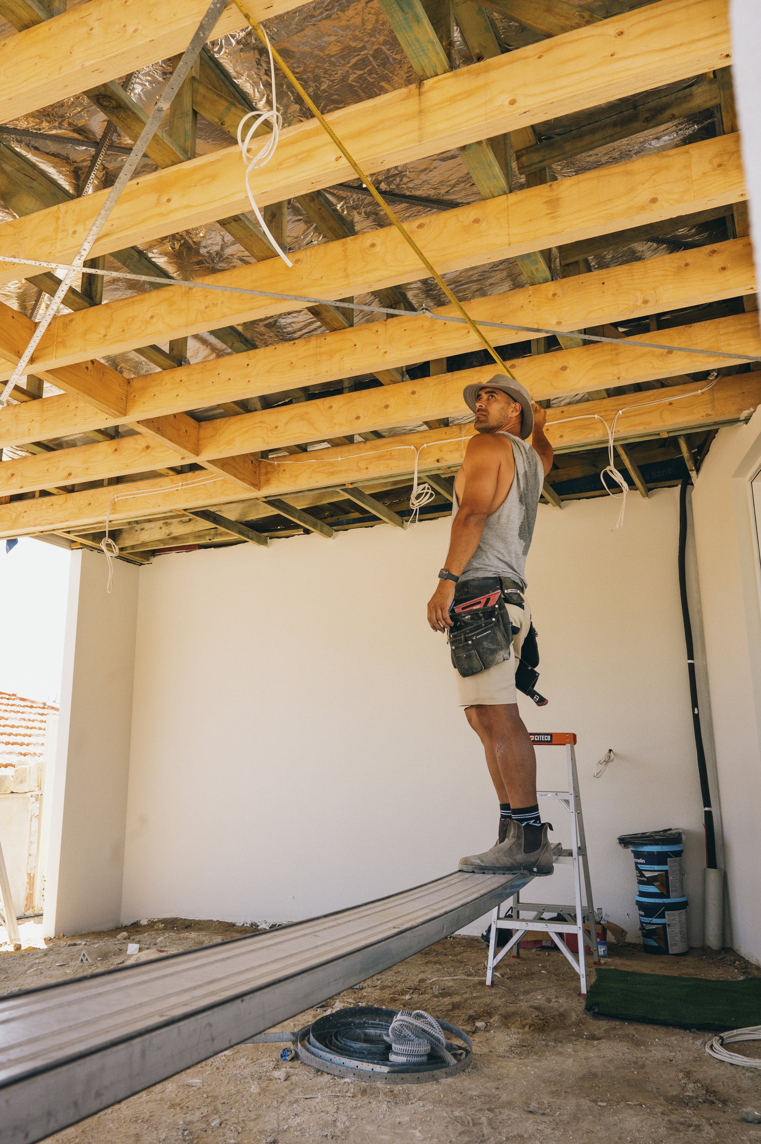 Construction worker balancing on a beam inside a building with exposed wooden ceiling joists and wiring.