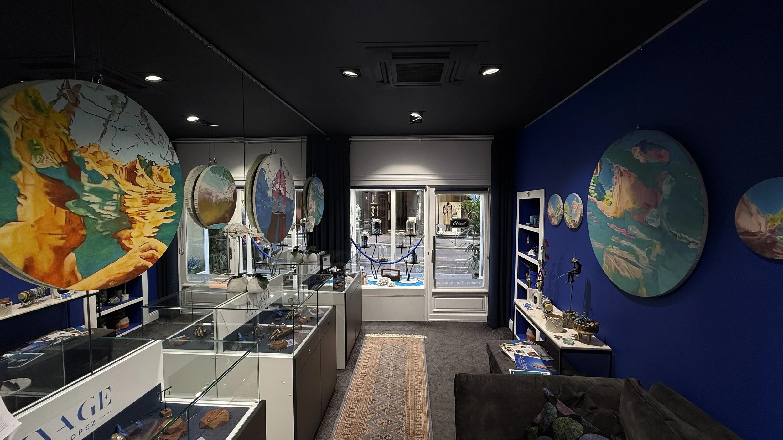 Intérieur d'une boutique d'art avec des peintures circulaires colorées sur un mur bleu, des vitrines avec des objets d'art et un tapis au sol.