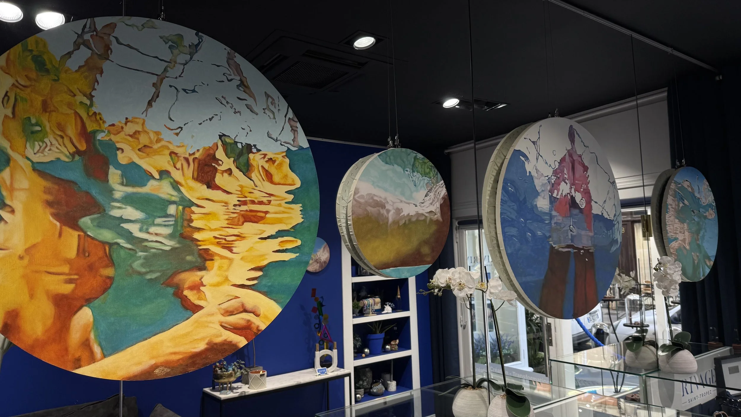Exposition de tableaux ronds suspendus avec des paysages colorés dans une salle d'art, décorée avec des vases et des fleurs blanches, fenêtres laissant entrer la lumière naturelle.