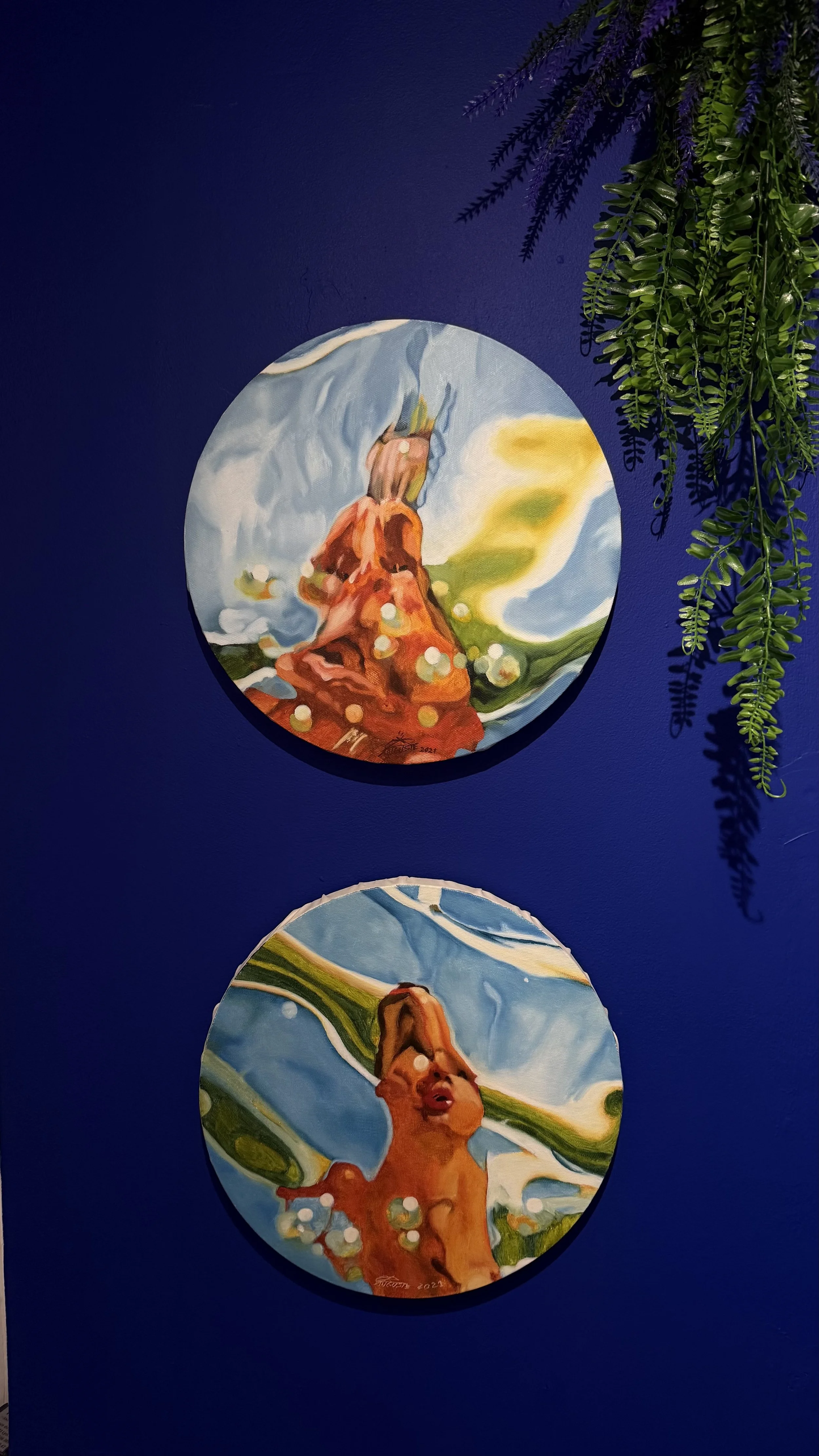 Deux tableaux ronds représentant une femme avec une coiffure en hauteur, dans un style artistique coloré. Le fond du mur est bleu foncé, avec des plantes vertes suspendues à droite.