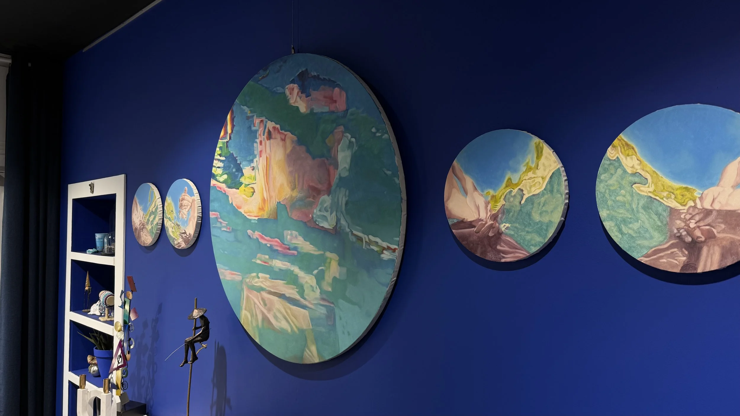 Quatre peintures rondes représentant des paysages abstraits avec des montagnes et des rivières, accrochées sur un mur bleu, avec une étagère blanche contenant des objets décoratifs à gauche.