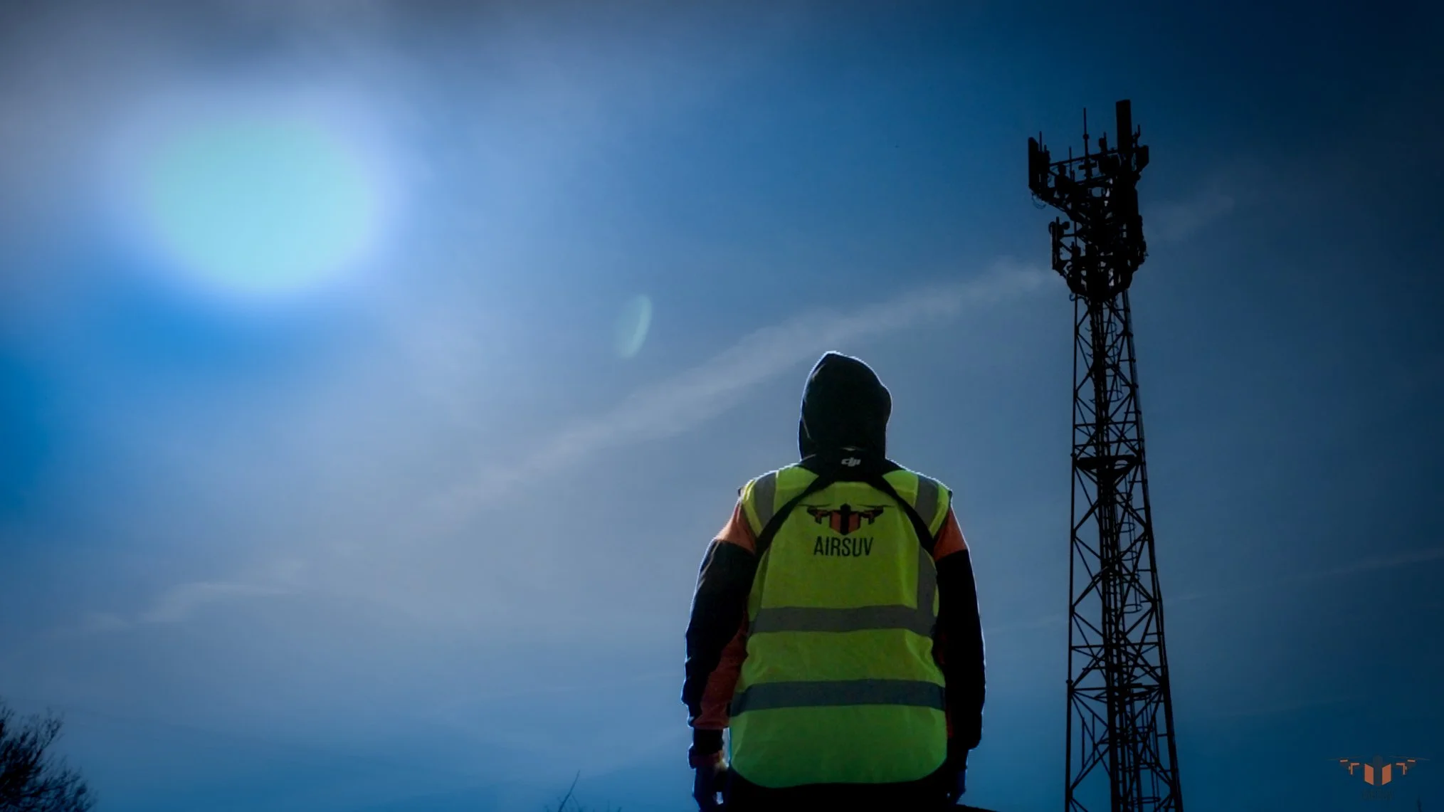 Person wearing a high-visibility vest labeled 'AIRSUV' standing outdoors facing a tall communication tower.
