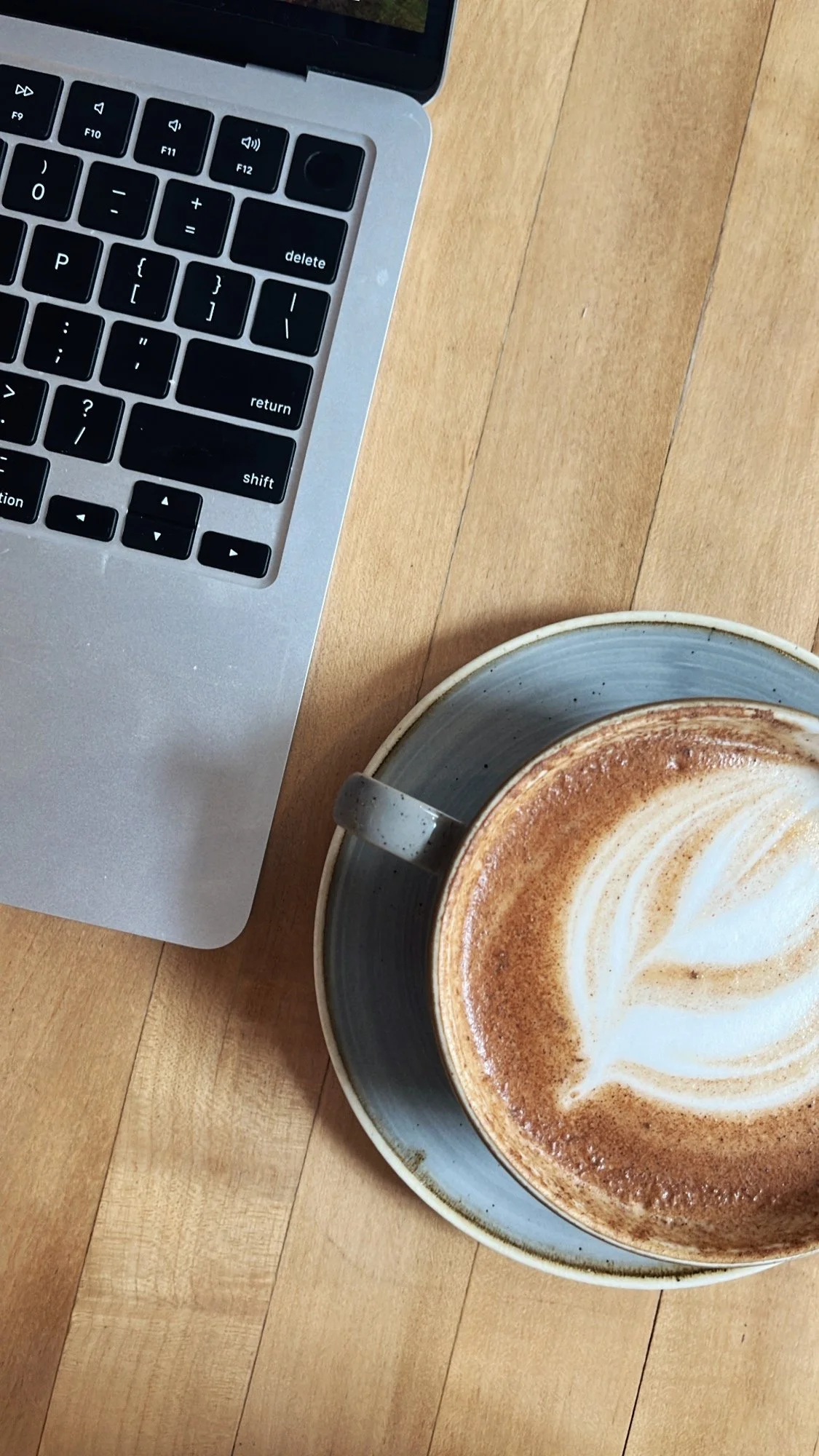 The keyboard of a silver laptop can be seen in the top left with a latte in a large mug is on the bottom right
