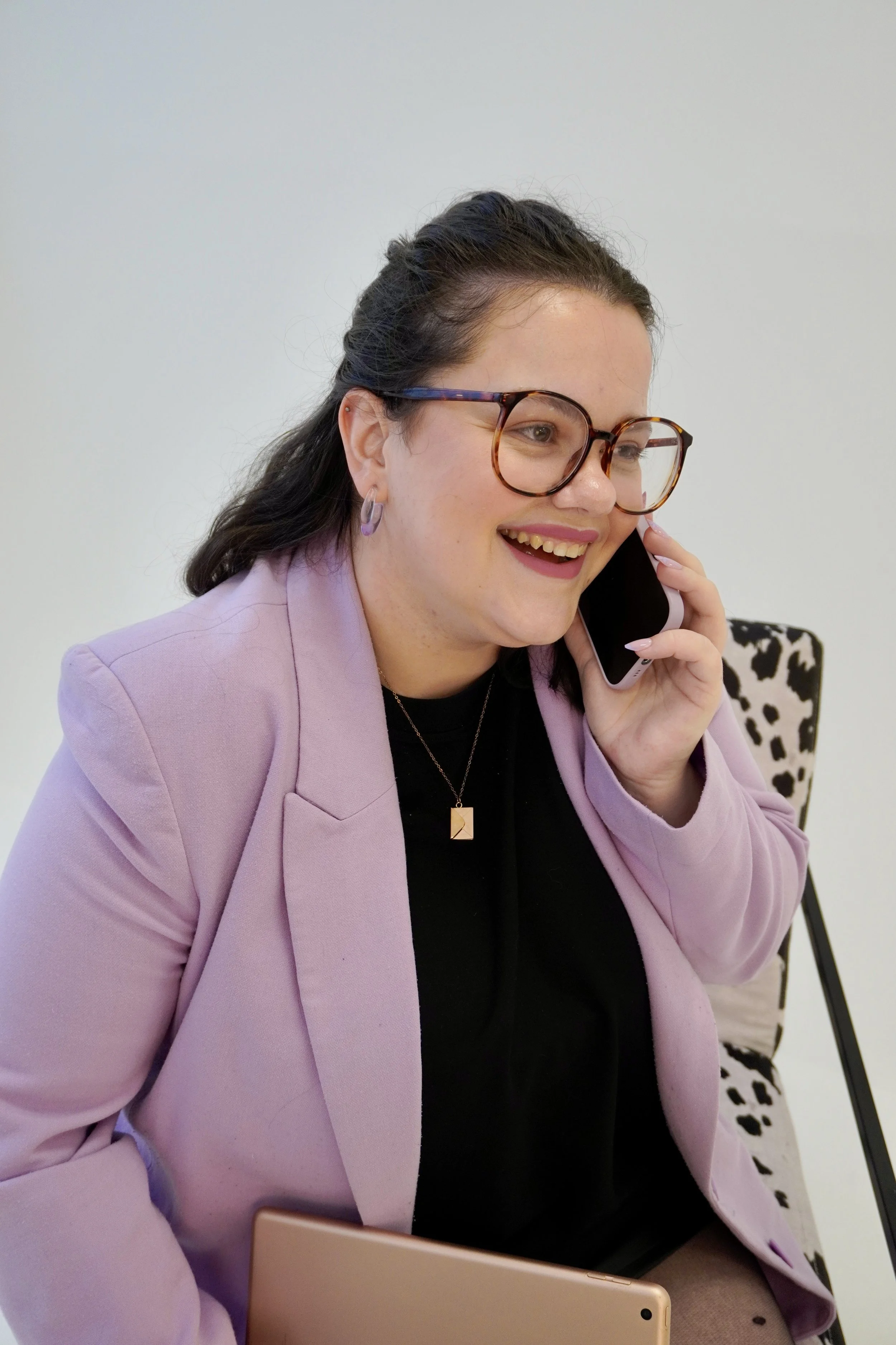 A woman with dark hair pulled behind her head is perched on a chair. She is wearing a black t-shirt and lilac blazer with a rose gold envelope necklace hanging down her chest. She is looking away from the camera while smiling talking on her phone