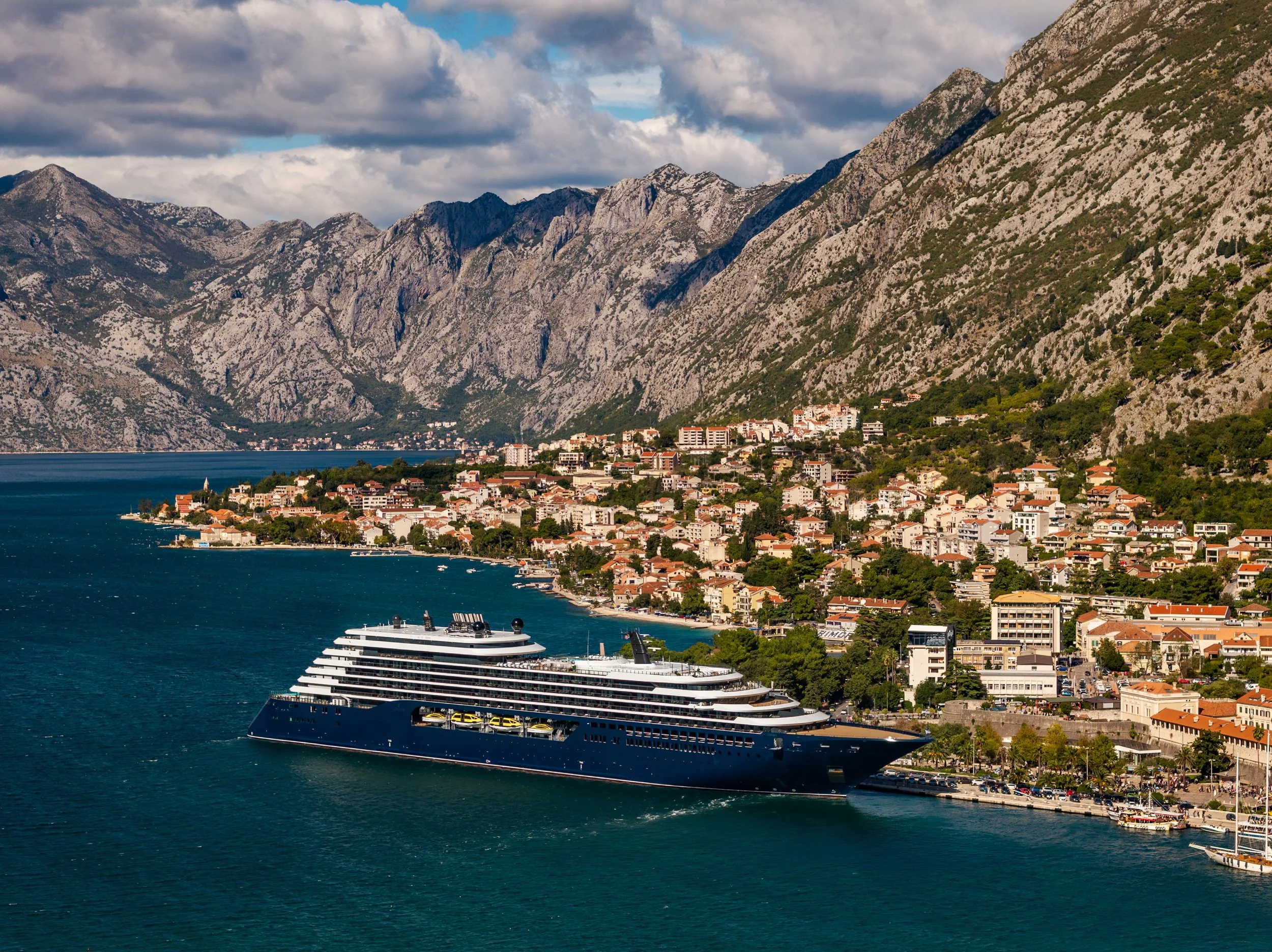 Ritz-Carlton Yacht Collection luxury cruise ship docked in the Bay of Kotor, Montenegro, surrounded by dramatic mountains and coastal town.
