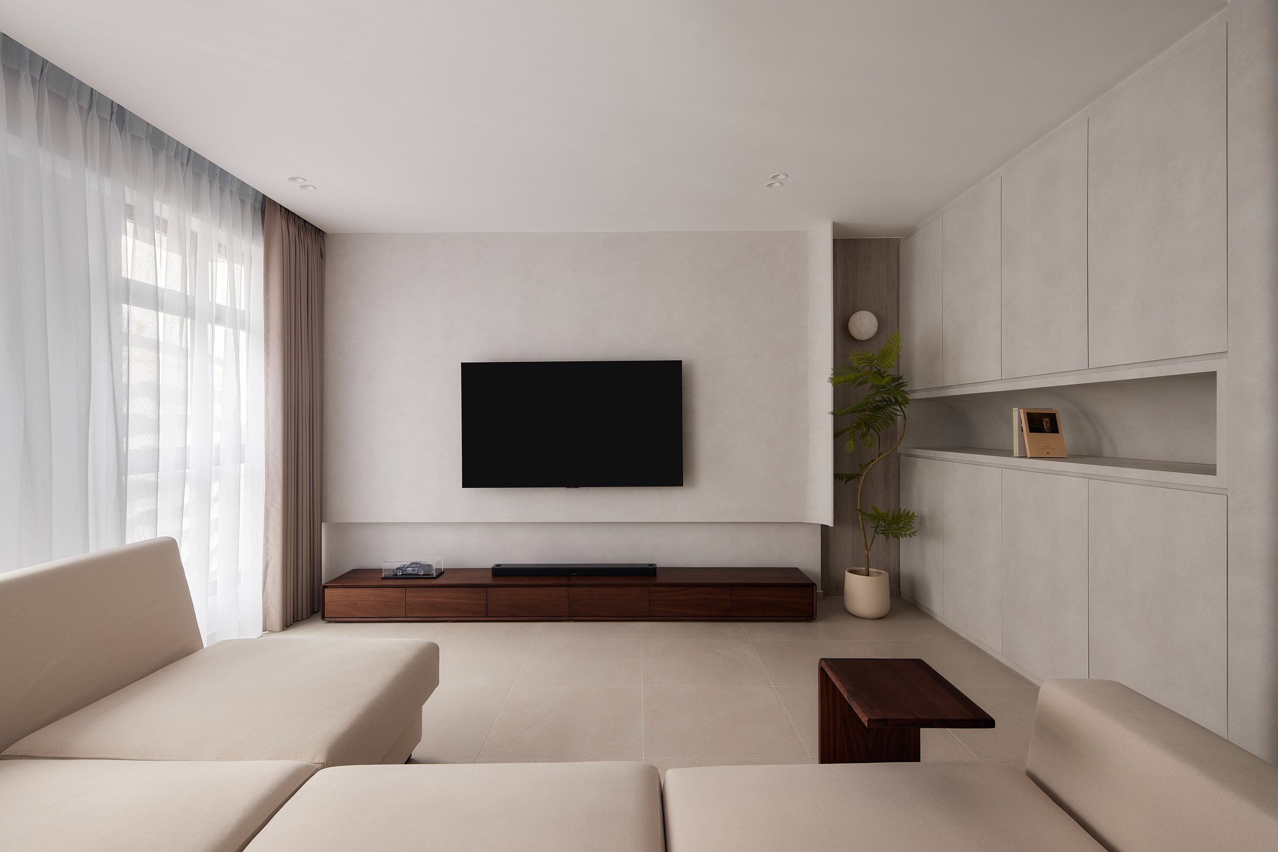 Modern living room with beige sofa, wall-mounted TV, dark wood media console and large window with sheer curtains.