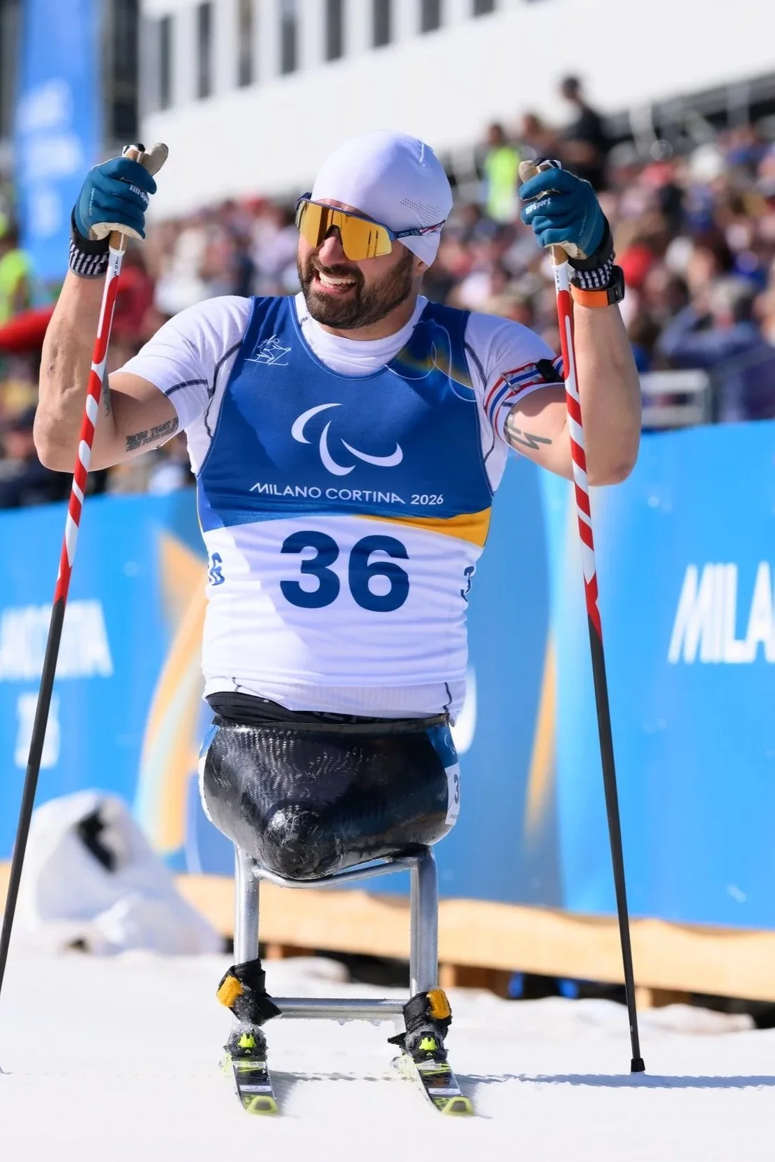Paralympic athlete sitting in a nordic sit ski, wearing sunglasses, a white cap, and a blue and white team uniform, sitting in pain after a race at the Milan Cortina 2026 Winter Paralympics.