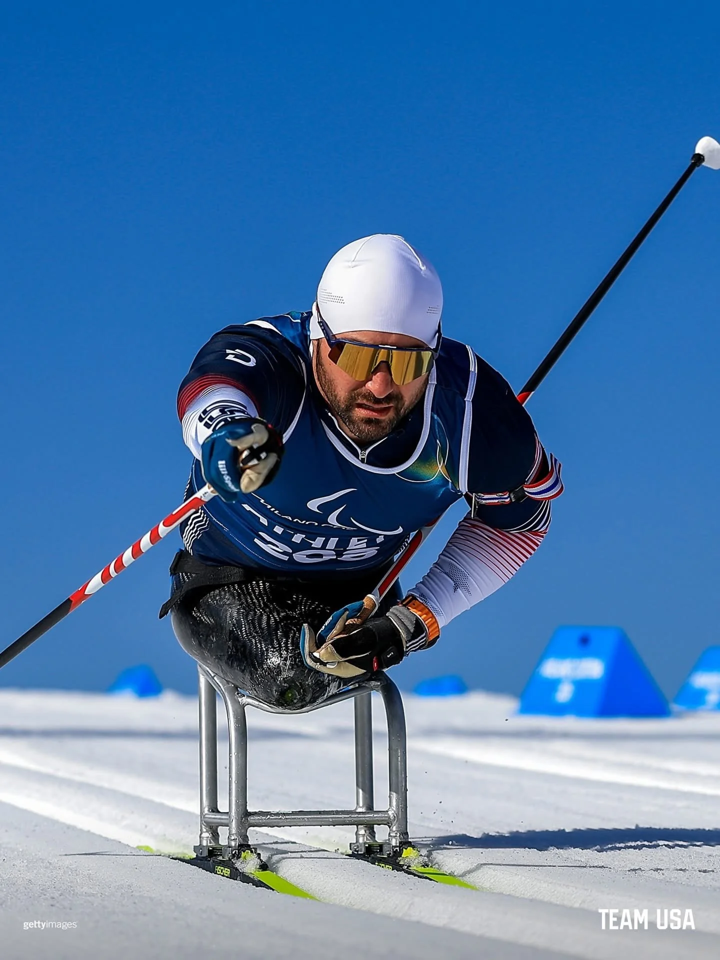Paralympic athlete racing on snow using a monoski and poles, wearing a white beanie and reflective sunglasses, with blue sky and blue course markers in background.