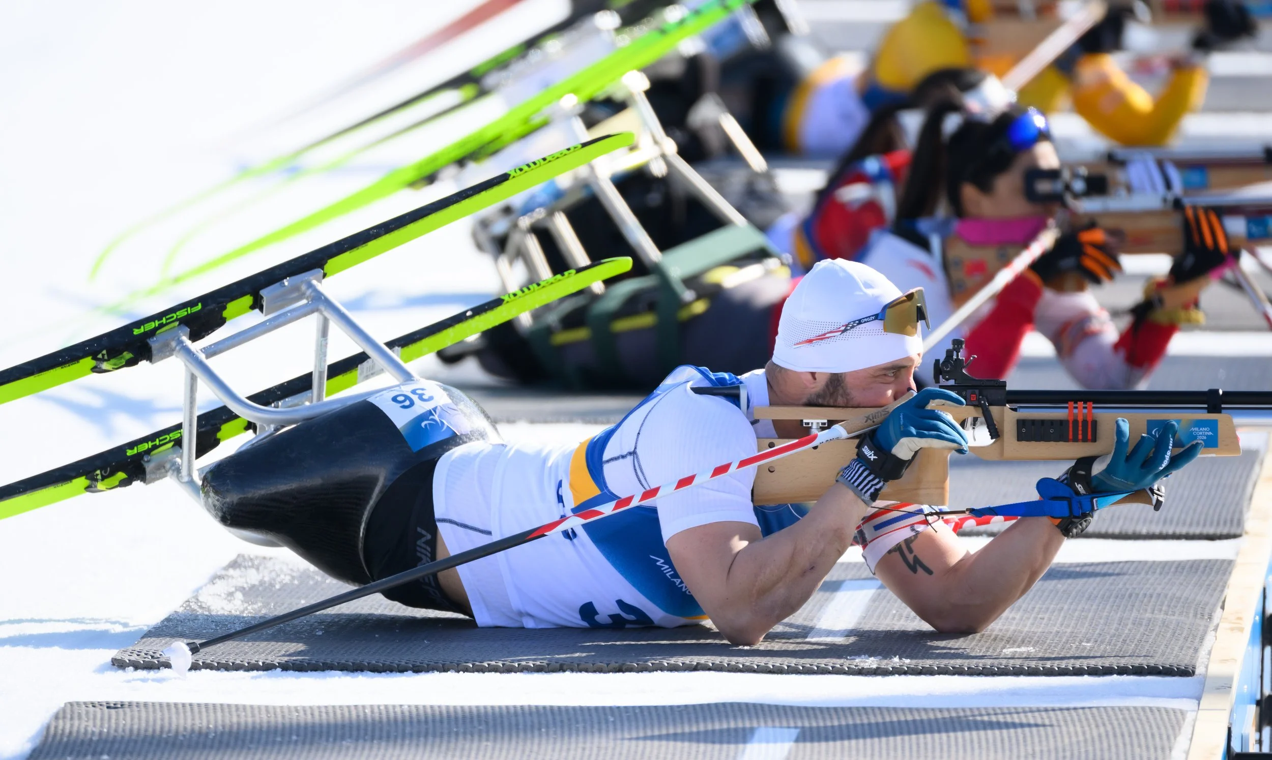 Biathlete aiming a rifle while lying on snow with others in the background.