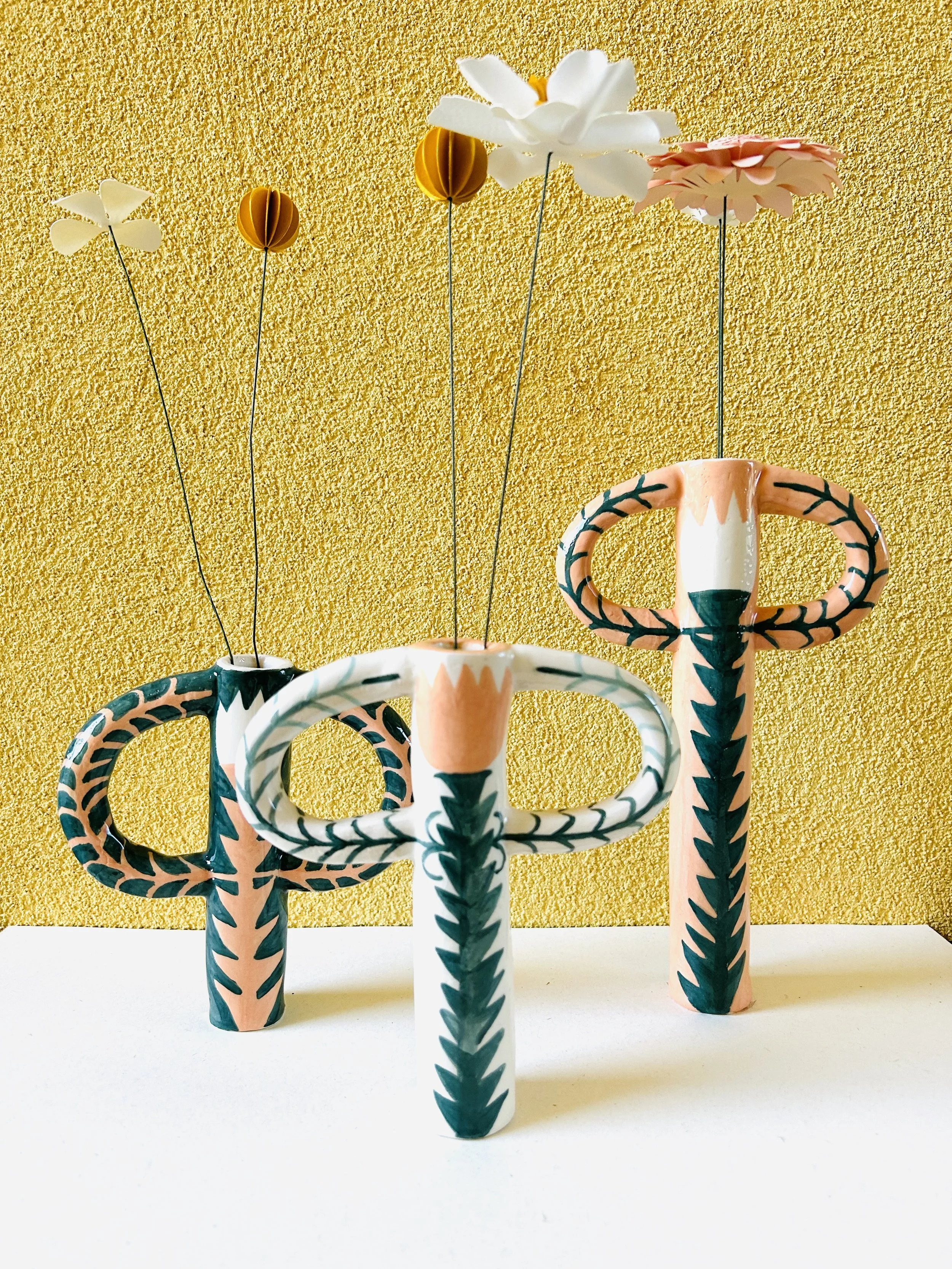 Tre ceramiche colorate con disegni geometrici e tre fiori di carta in vasi differenti, su una superficie bianca con sfondo giallo.