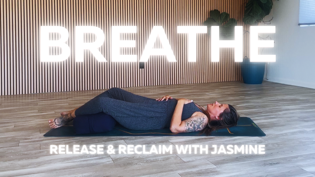 A woman lying on her back on a yoga mat with a bolster under her knees, practicing breathing exercises in a studio with a wooden wall and plant. The text 'BREATHE' is displayed prominently, along with 'RELEASE & RECLAIM WITH JASMINE' at the bottom.
