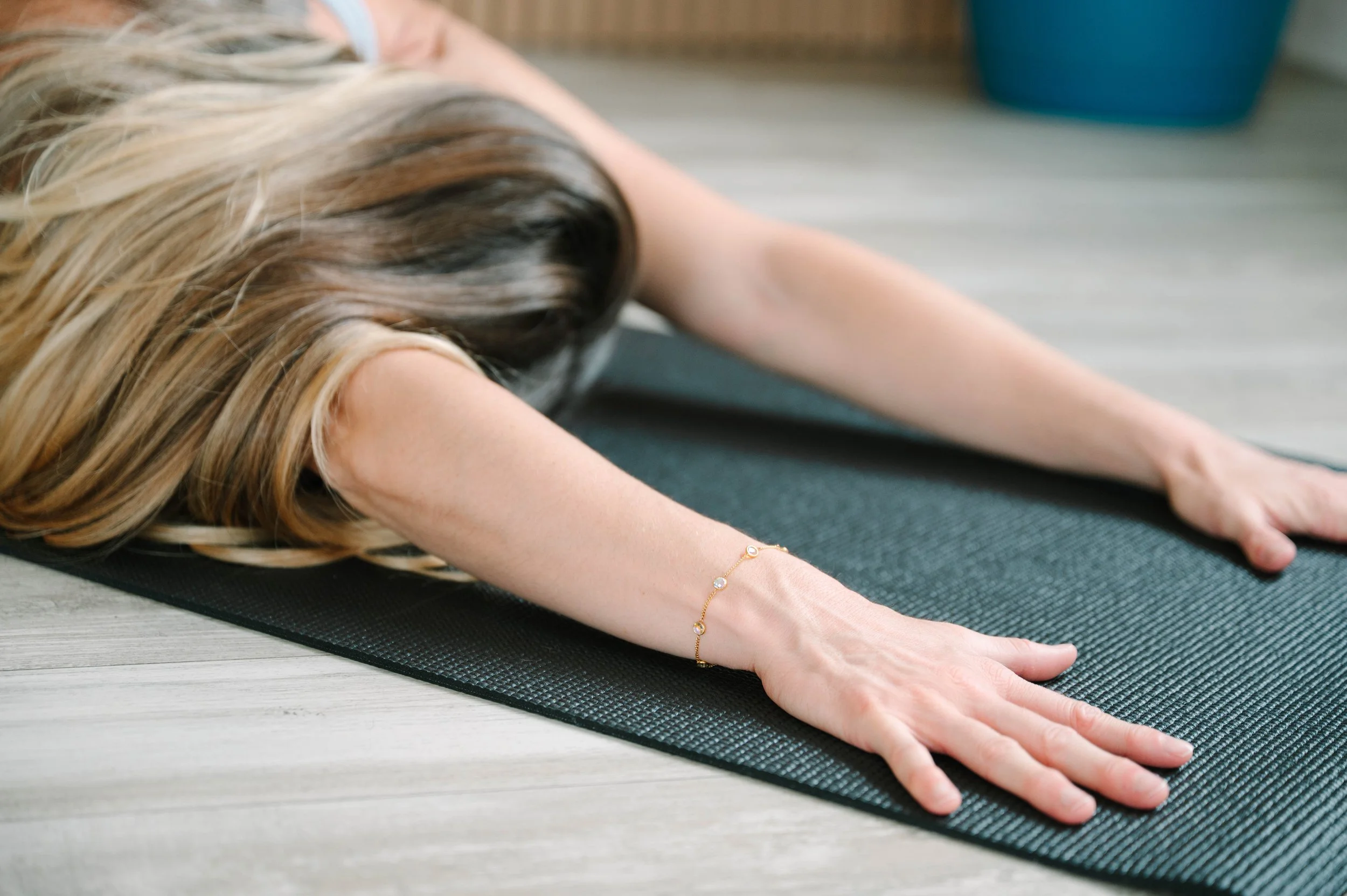 A woman with blonde hair practicing yoga on a black mat indoors, extending her arms forward, wearing a gold bracelet with small charms.