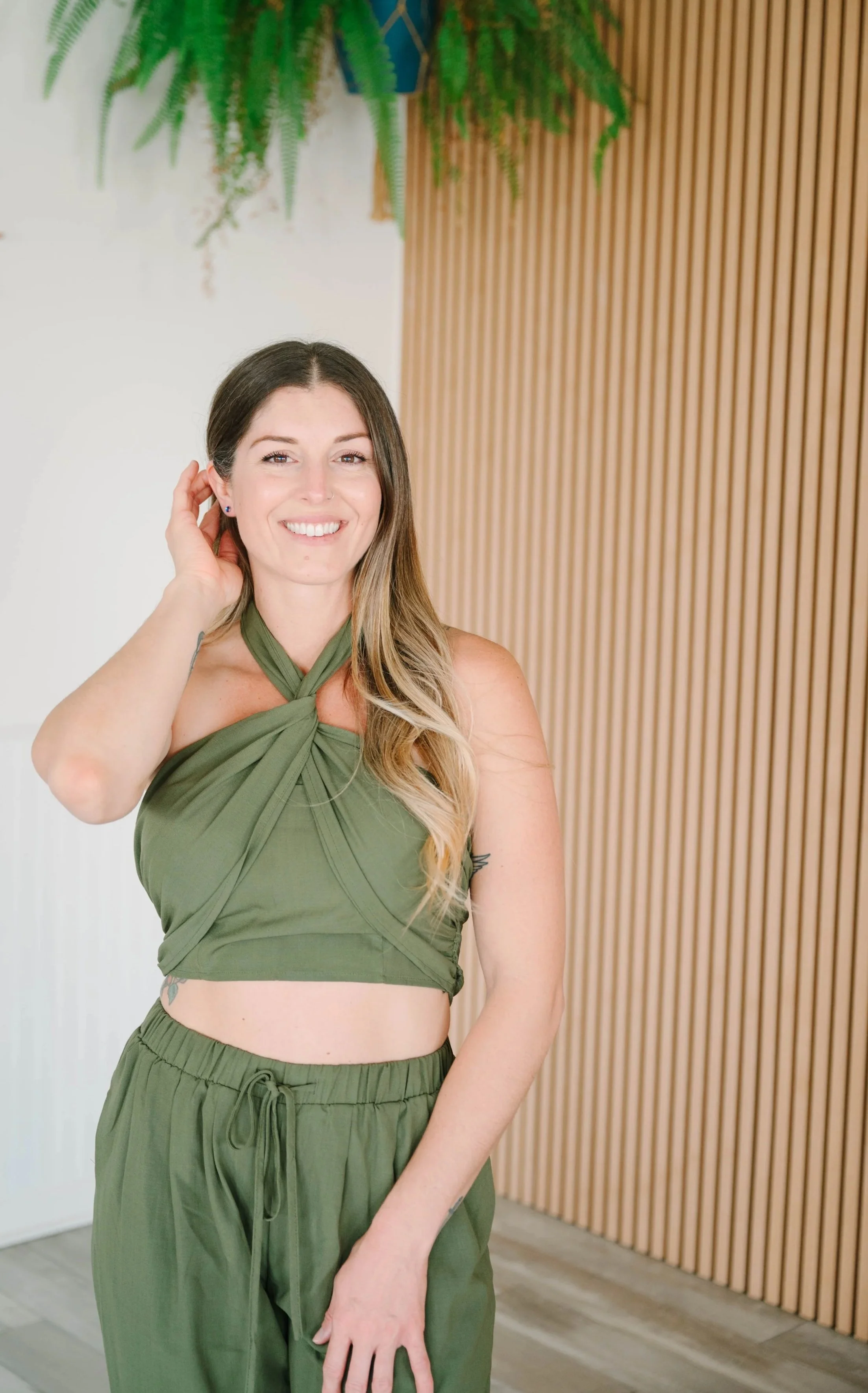 A woman smiling in a green halter top and matching pants, standing indoors near a wooden slat wall and greenery.