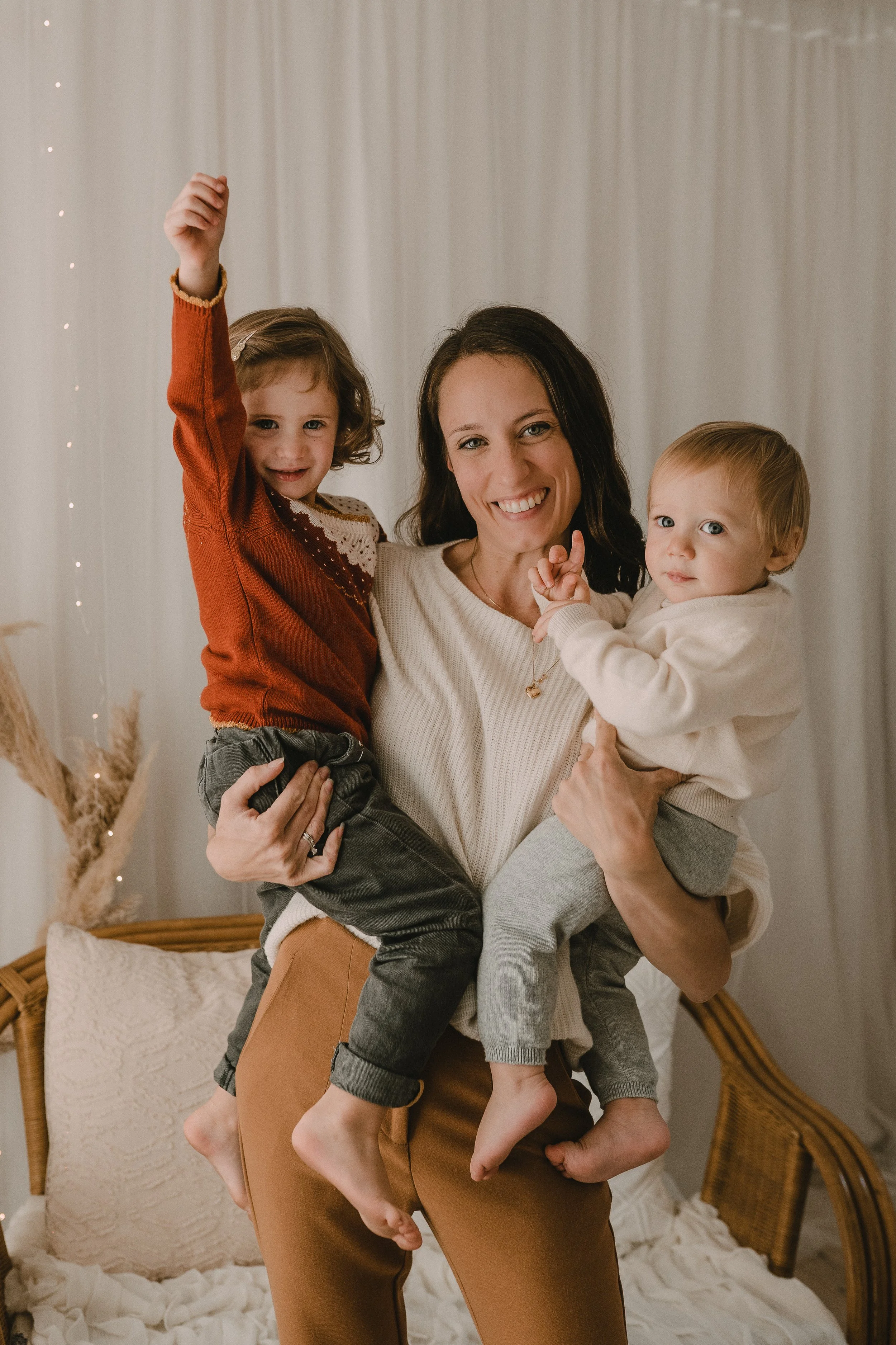 Una donna con due bambini, uno a sinistra e l'altro a destra, sorridenti e felici in un ambiente domestico, con sfondo di tende e decorazioni natalizie.