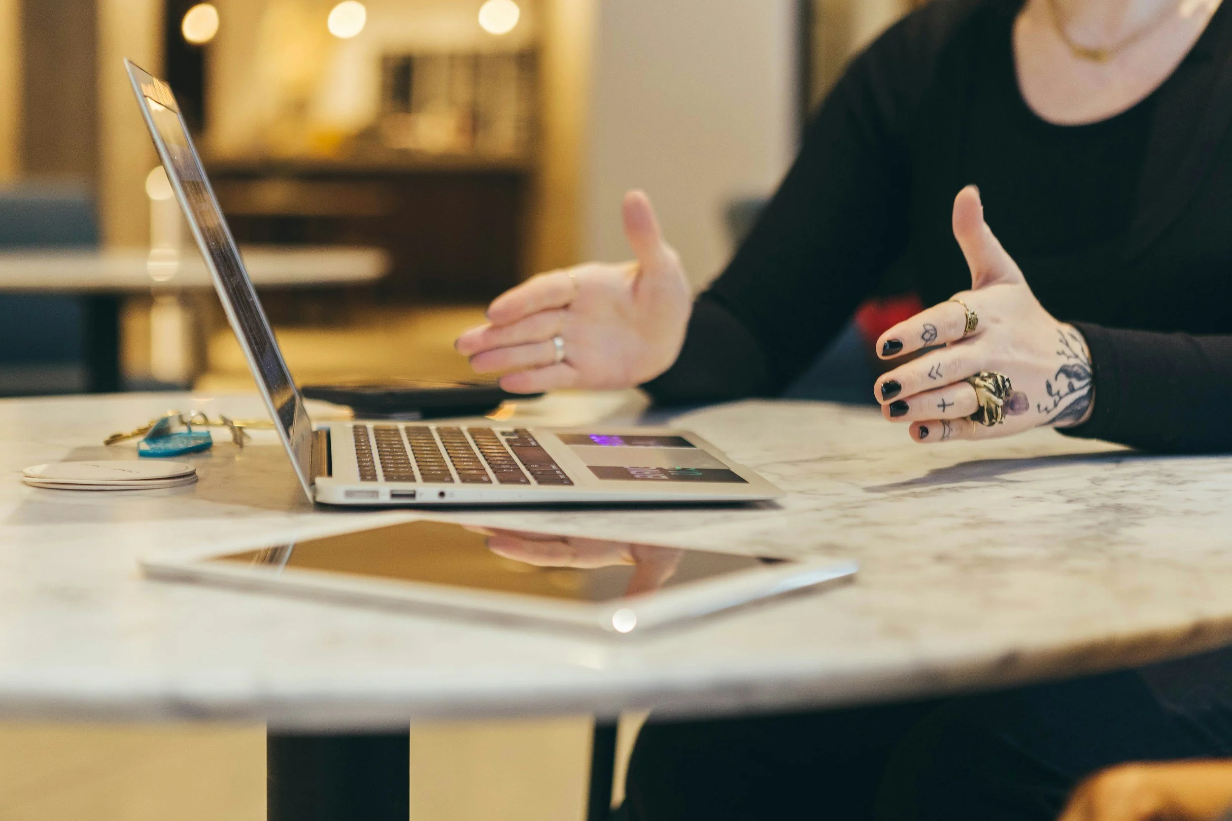 Person with tattoos and rings gesturing while sitting at a marble table with a laptop, keys, and a phone.