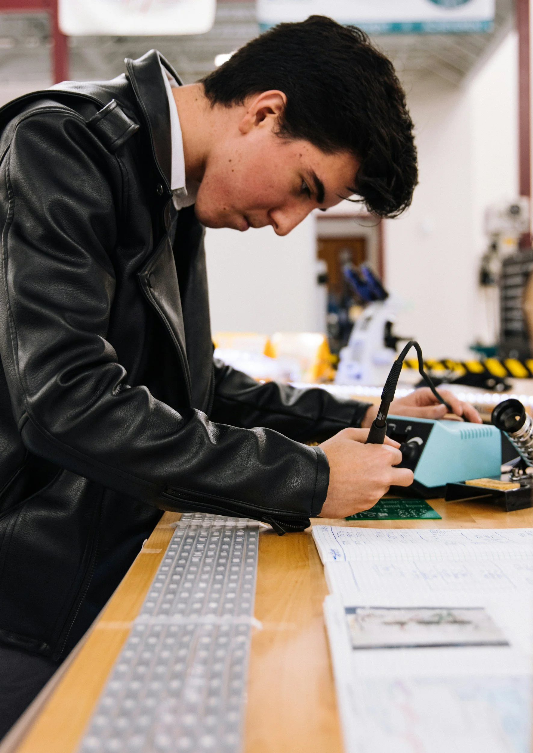 Young man working with electronic equipment and tools at a woodworking or electronics workshop, wearing a black leather jacket.